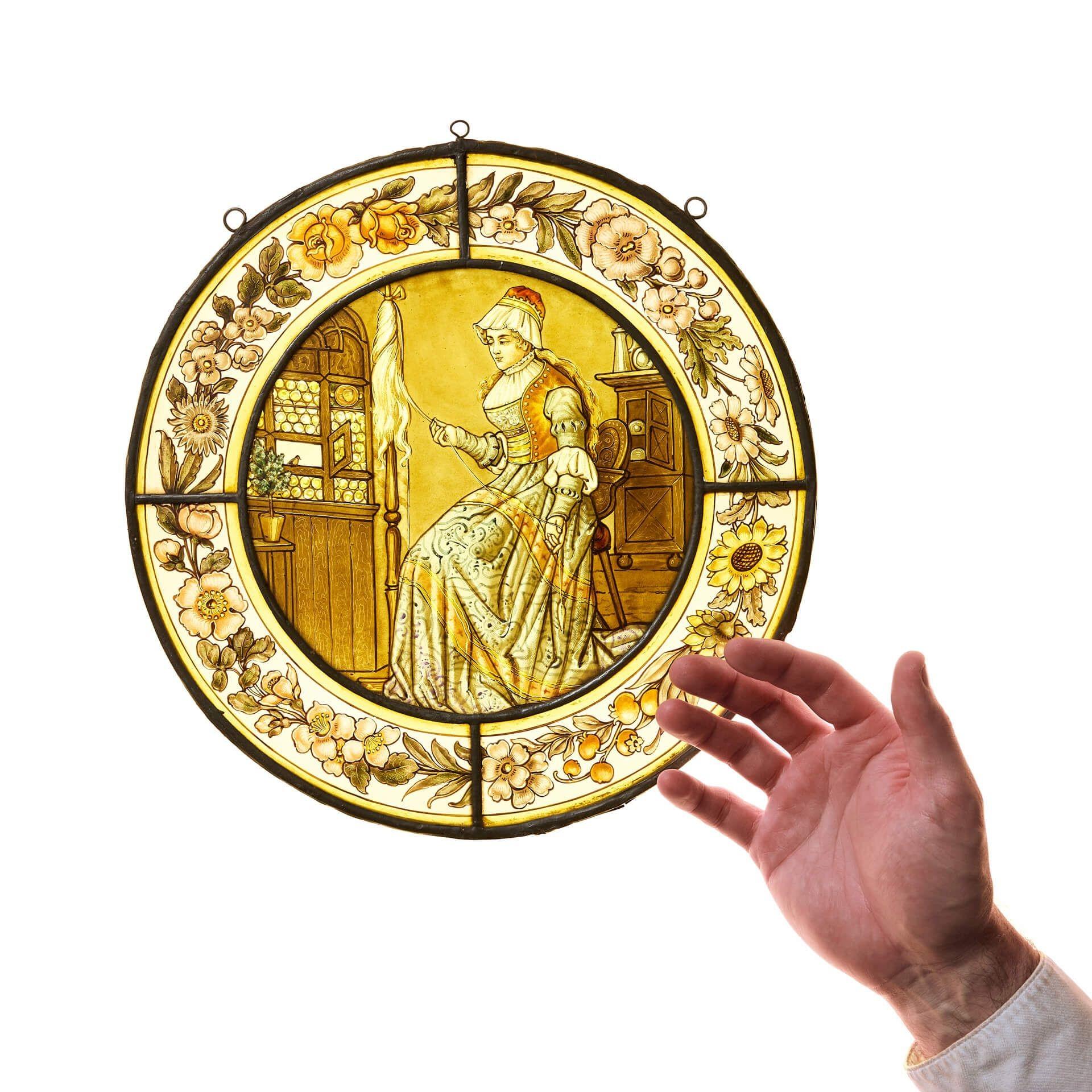 Un vitrail ancien représentant une femme victorienne en train de filer. Des fleurs stylisées peintes à la main et une bordure jaune vibrante entourent la femme pendant qu'elle file le fil sans effort. Sa robe est d'un bleu pâle et d'or, ce qui