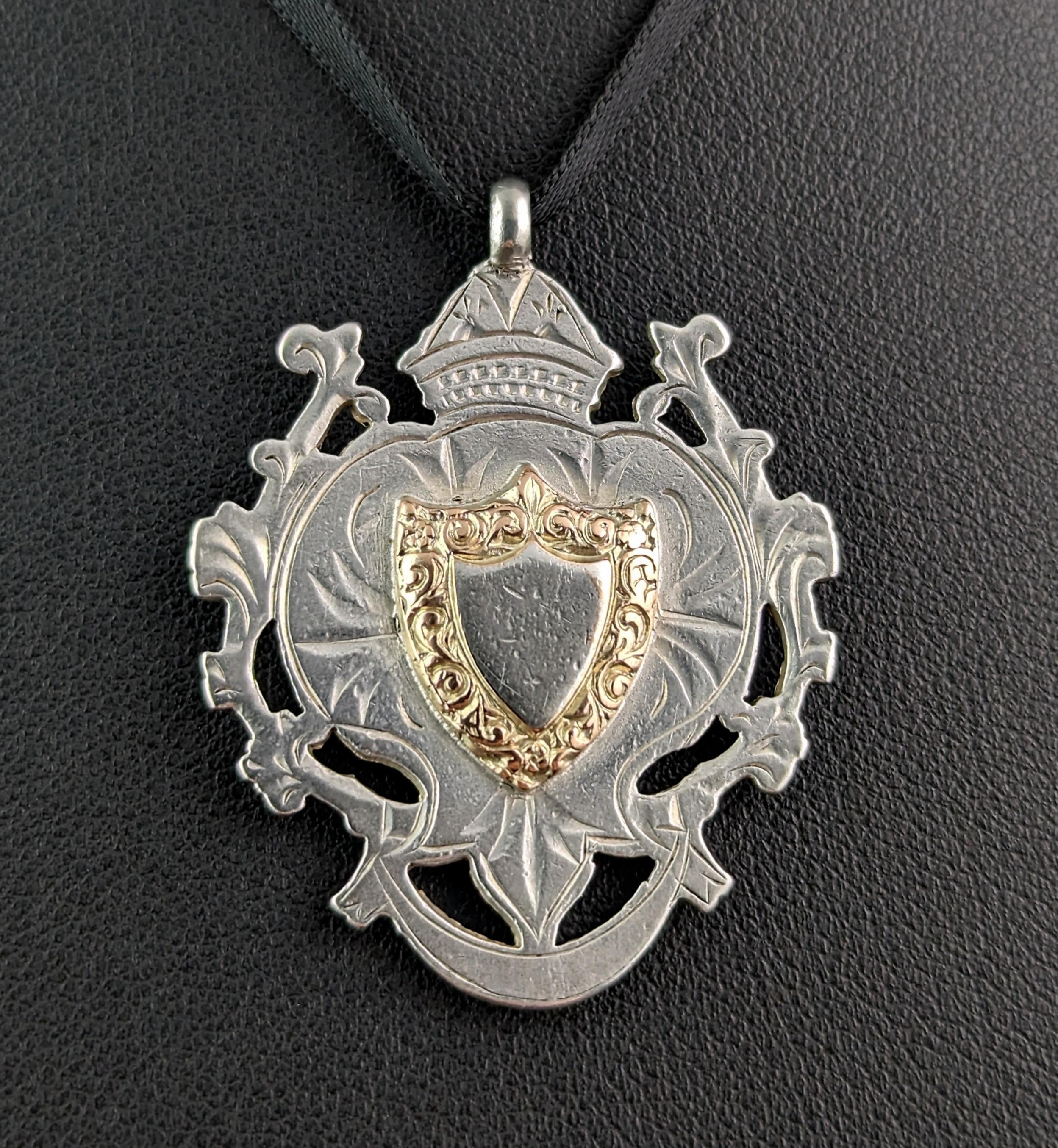A lovely antique Edwardian era sterling silver and 9ct gold watch fob pendant.

It is an almost heart shaped piece with a scrolling border and is crowned to the top, to the centre there is a small shield shaped cartouche with a chased and engraved