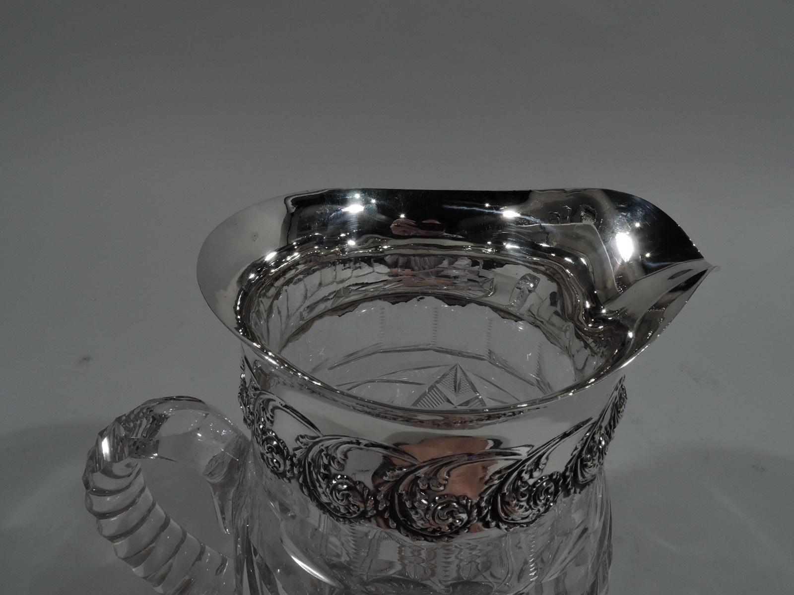 Edwardian cut-glass water pitcher with sterling silver collar. Curved body with cut geometric and plant ornament and faceted c-scroll handle. Collar has band of volute scrolls with flowers and leaves. Fully marked including stamp for Adelphi, a New