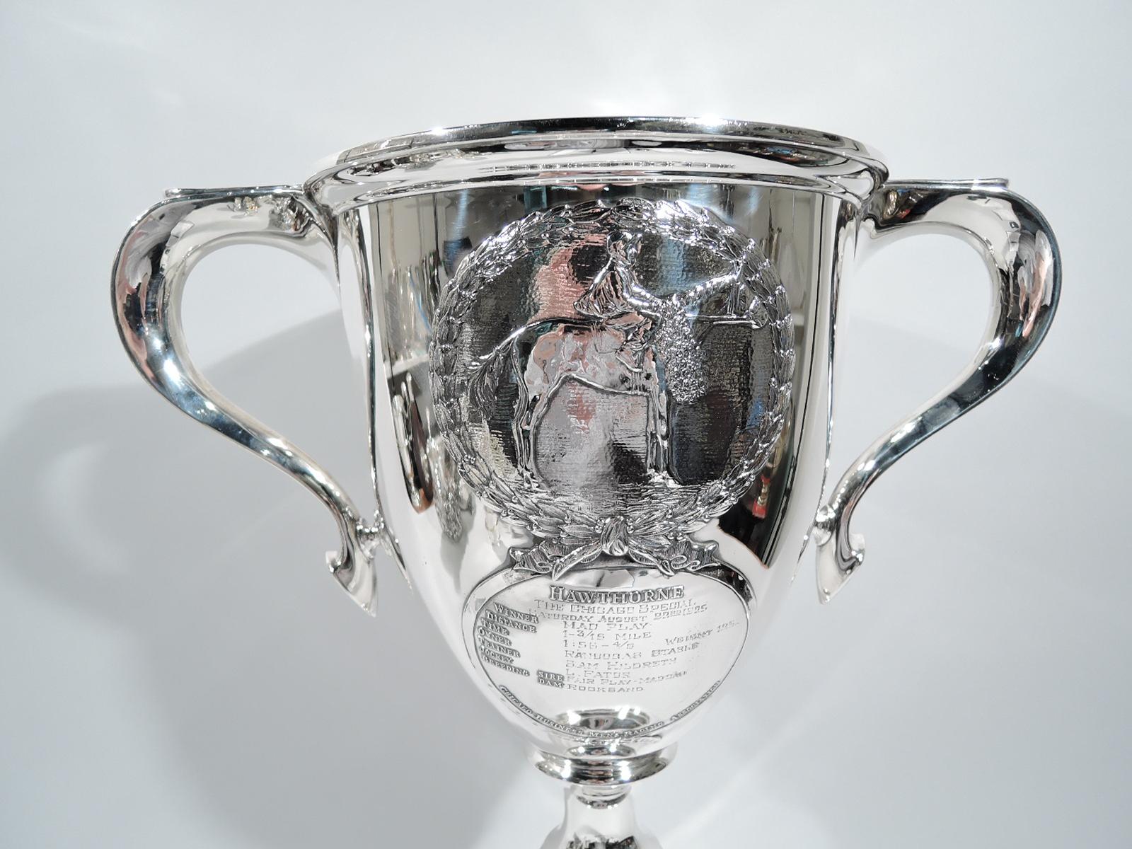 Edwardian sterling silver trophy cup. Oval bowl with capped and scrolled side handles; spool support and raised and stepped foot. On front is applied is a wreath with mounted horse covered in laurels. Below is a round strap frame with “Hawthorne” at