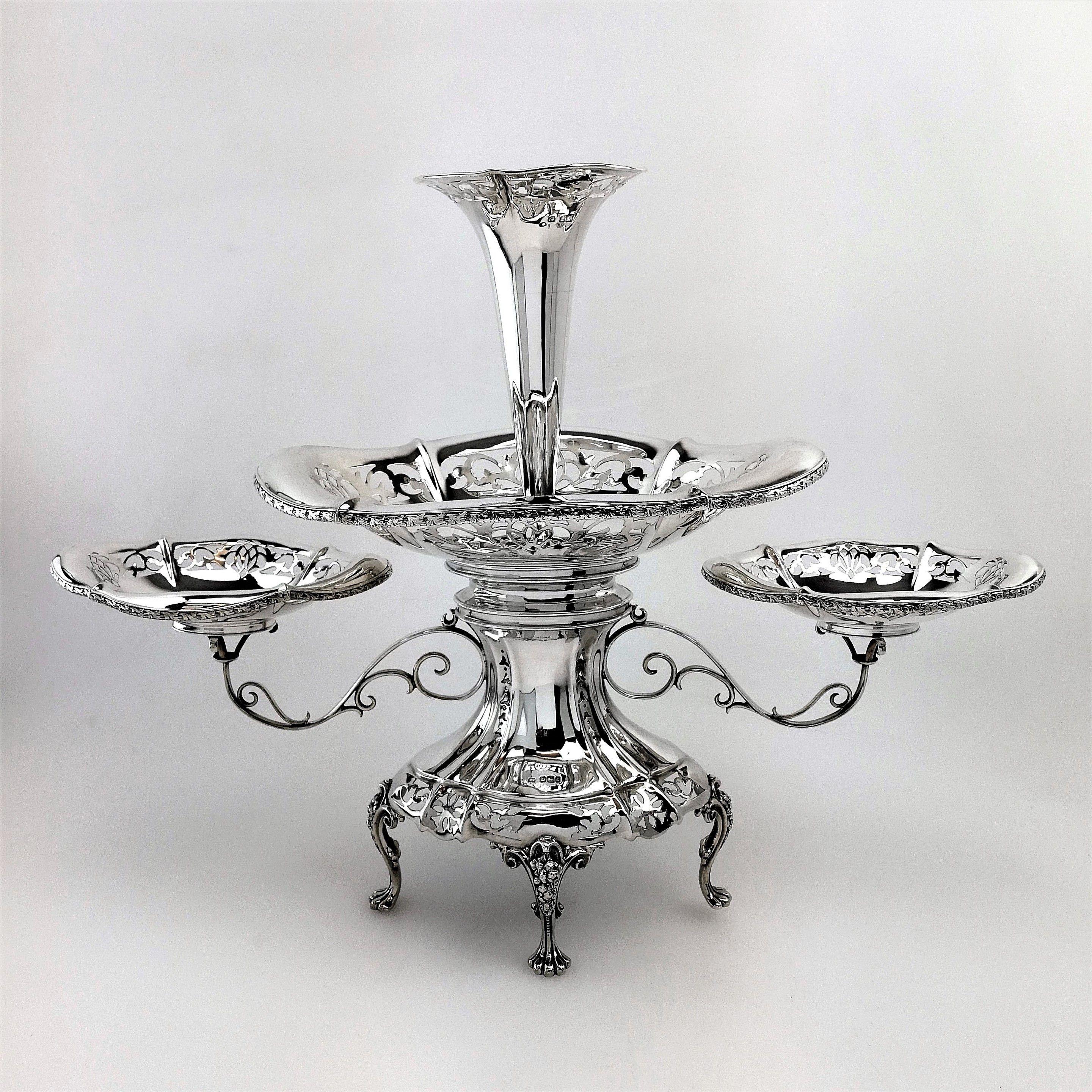An Antique solid Silver Epergne Centrepiece with a large oval central basket supporting a tall trumpet shaped vase on top of an impressive concave shaped stand. The central stand has two scrolled branches supporting a matched pair of smaller oval
