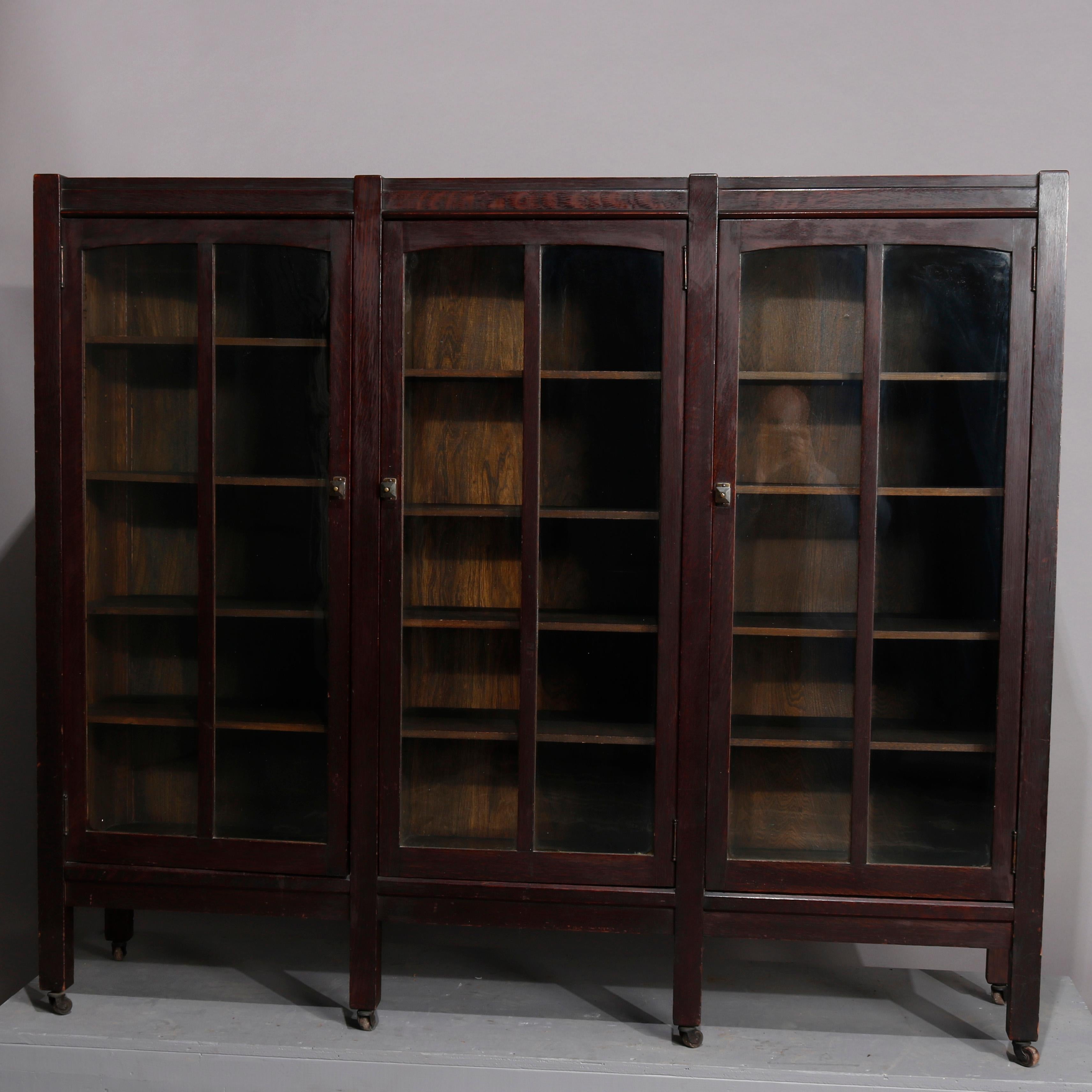 An antique Stickley School Arts & Crafts mission oak bookcase offers quarter sawn oak construction with three glass doors having arched frames and opening to shelved interior, raised on square and straight legs with casters, circa 1910

Measures:
