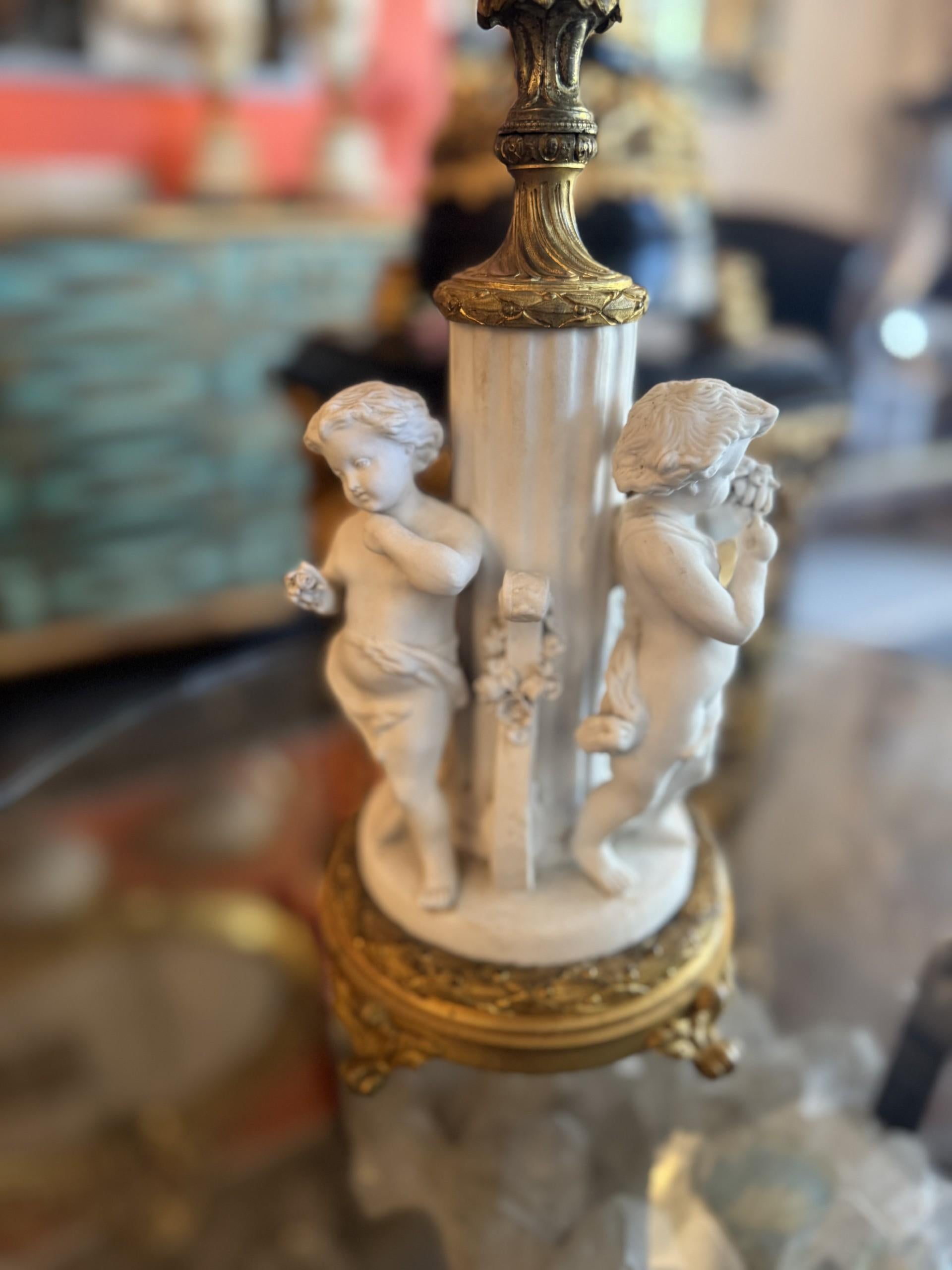 This elegant table lamp features a beautiful, detailed base with three classical cherub figures sculpted in a white marble-like finish. The ornate gold accents and fluted column add a touch of opulence, while the soft cream shade provides a warm,