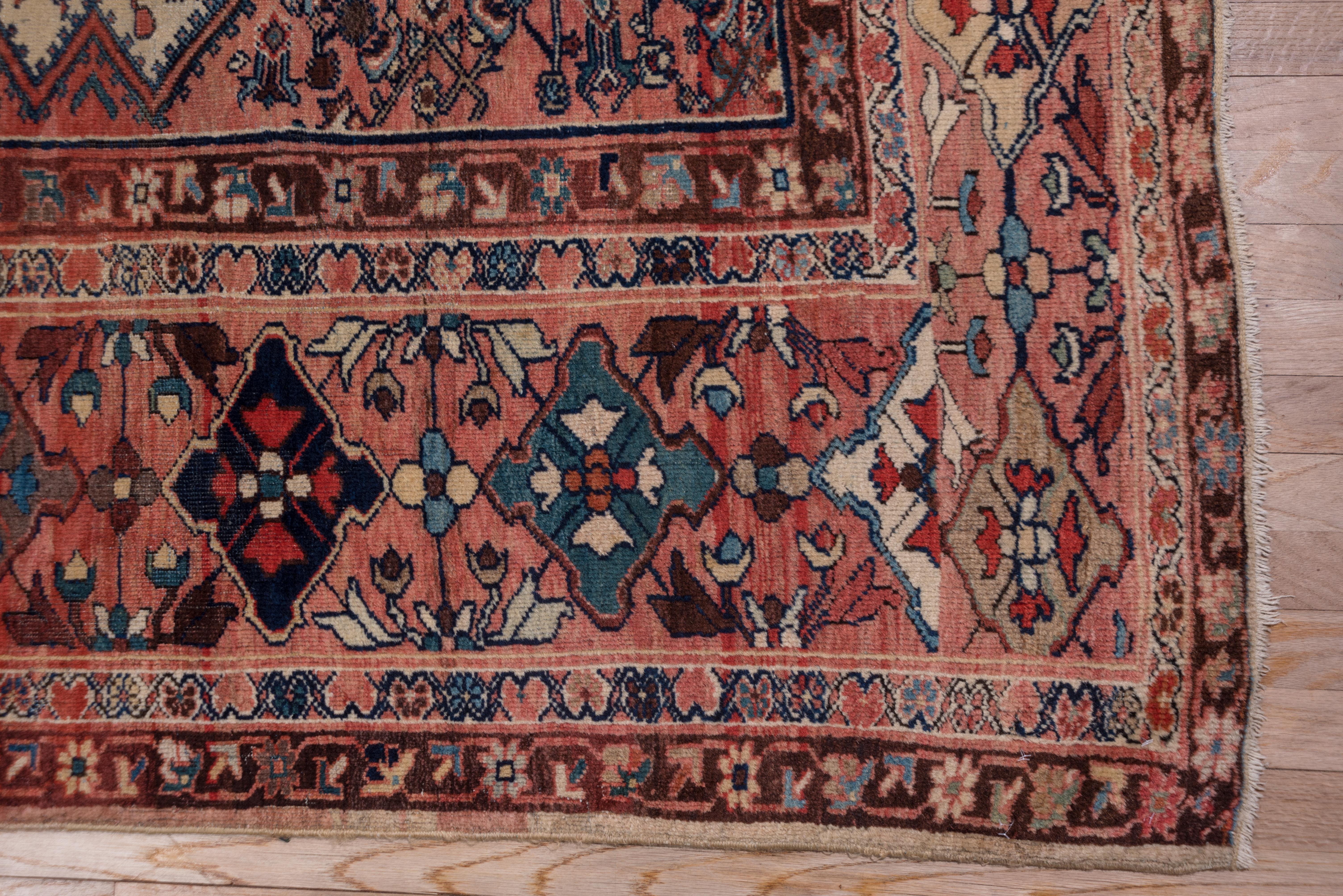 Ein sanftes rotes Rautenmedaillon mit gestuftem Umriss und Herati-Inhalt ist auf ein altes Elfenbeinfeld mit einem ebenfalls gestuften All-Over-Herati gesetzt. Die korallenfarbene, lachsfarbene Hauptbordüre zeigt gekerbte Rauten, die ein Blumenkreuz