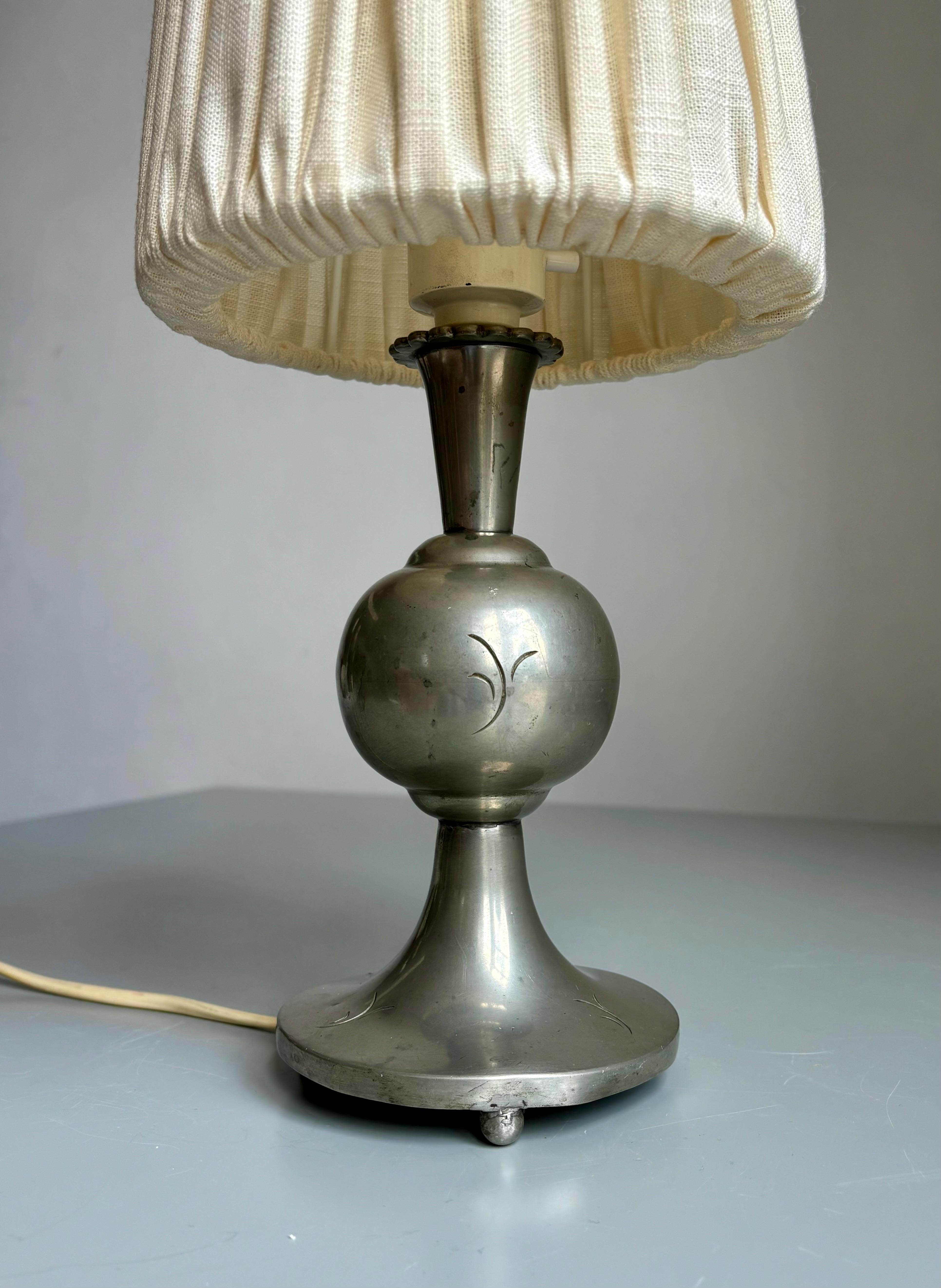 Antique pewter table lamp with floral decorations. Three spherical legs keeping it stable and lifted from the surface. Flower collar under fitting for E14 bulb. Small relief decorations on the base and globe shaped body under the slender neck.