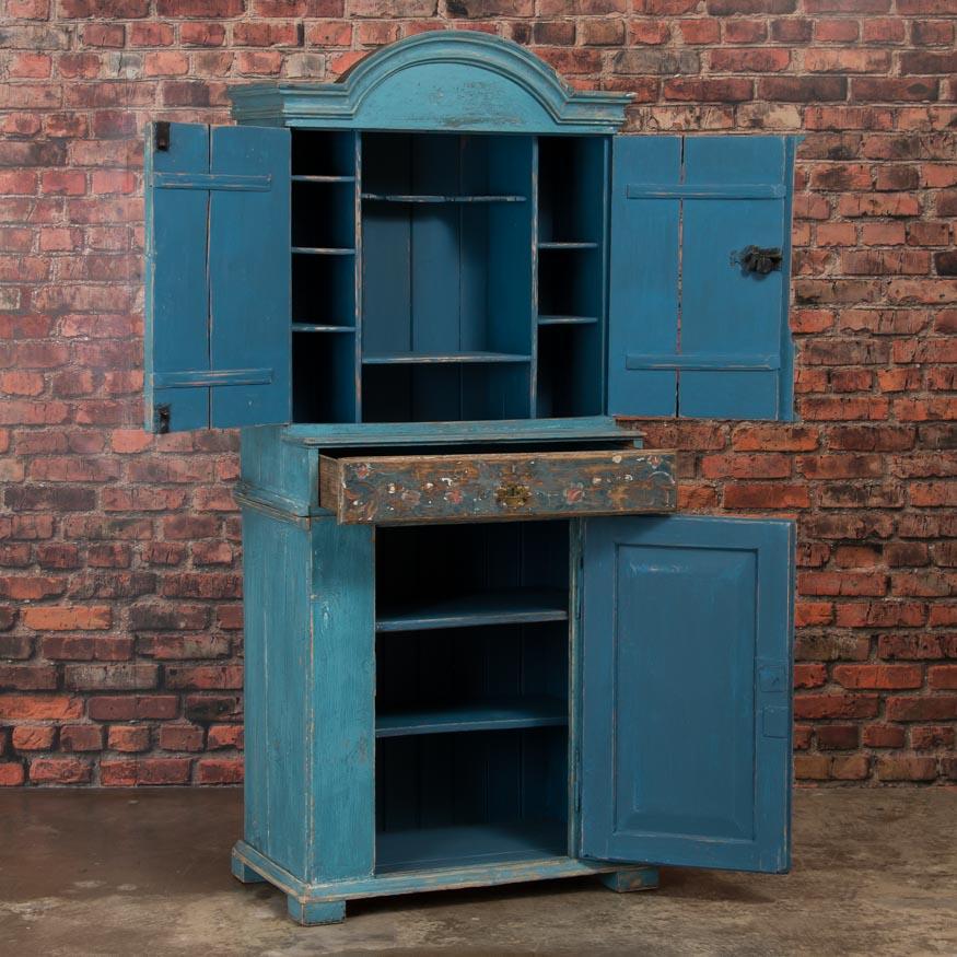 This style of cupboard with an arched molded crown, was traditionally painted in earth tones, which makes the original shade of blue on this exceptional cabinet unique. The doors and drawer are painted in a floral motif - the traditional folk art