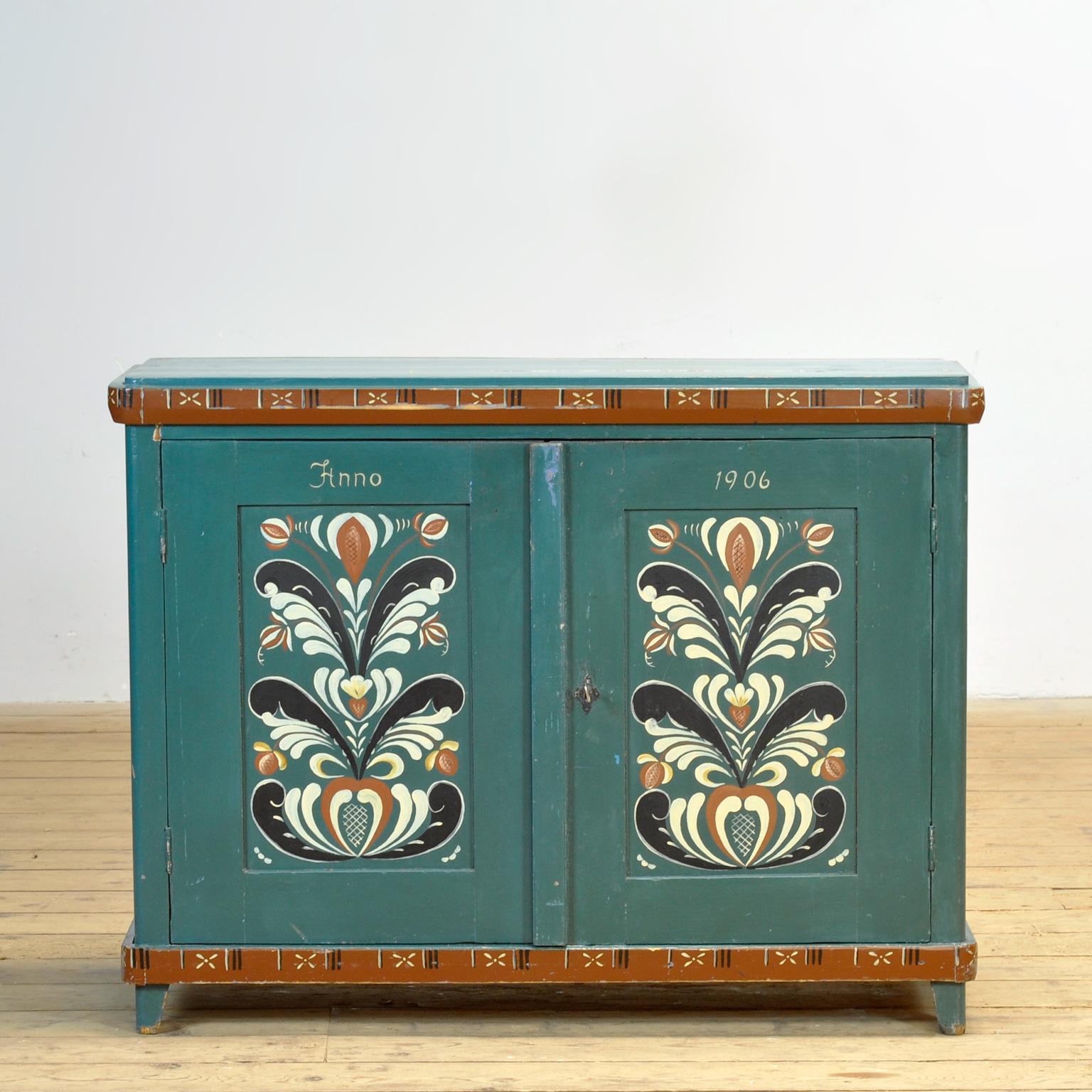 Les portes à deux panneaux sont peintes d'un motif floral traditionnel sur un fond bleu-vert. Ce meuble est une charmante représentation de l'artisanat suédois du début du XXe siècle, tant par l'ébéniste que par le peintre qui l'a créé. Veuillez