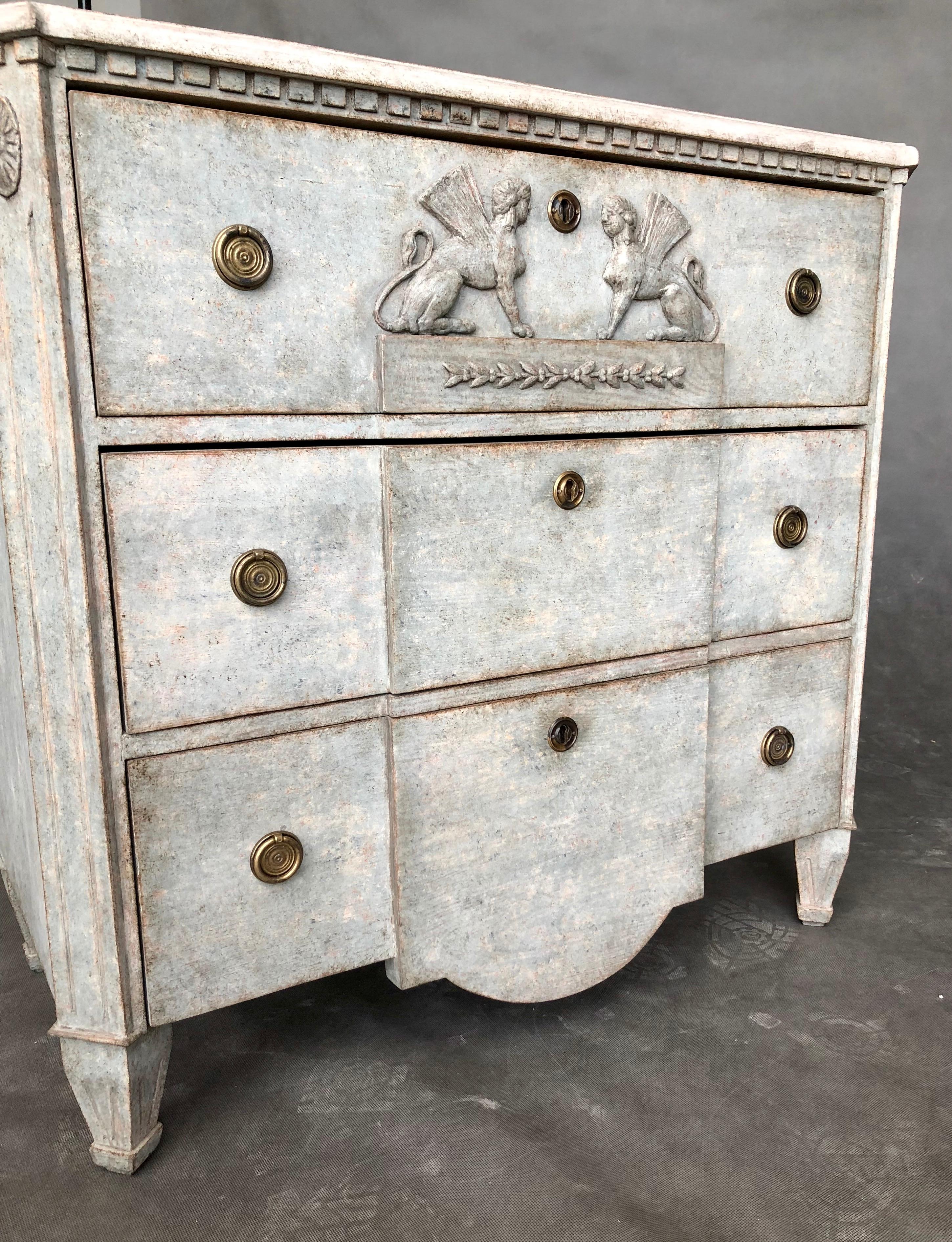 This is a neoclassical chest of drawers with a break front in gray color. The upper part of the furniture is decorated teeth. The corners are cut and there are vertical cutters and decorations on them. On the top drawer are sphinxes and subtle