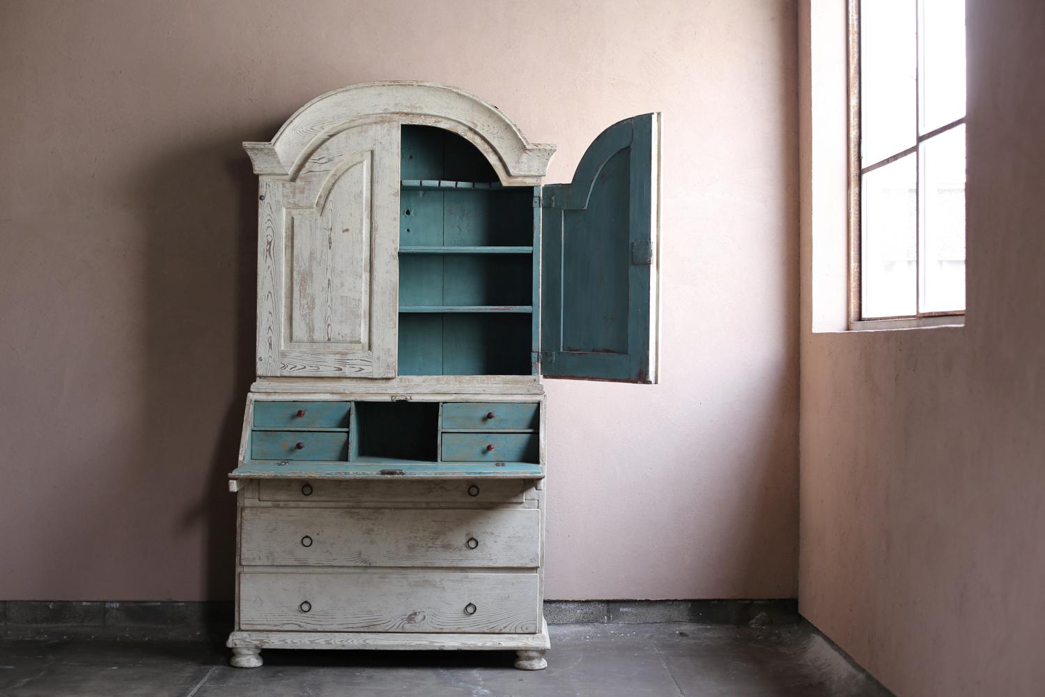 Questo antico mobile dipinto proveniente dalla Svezia risale al XIX secolo. Mentre l'elegante corona ad arco e le decorazioni delle porte ricurve mostrano influenze dello stile French Rococo, l'esterno dipinto di bianco e la silhouette slanciata
