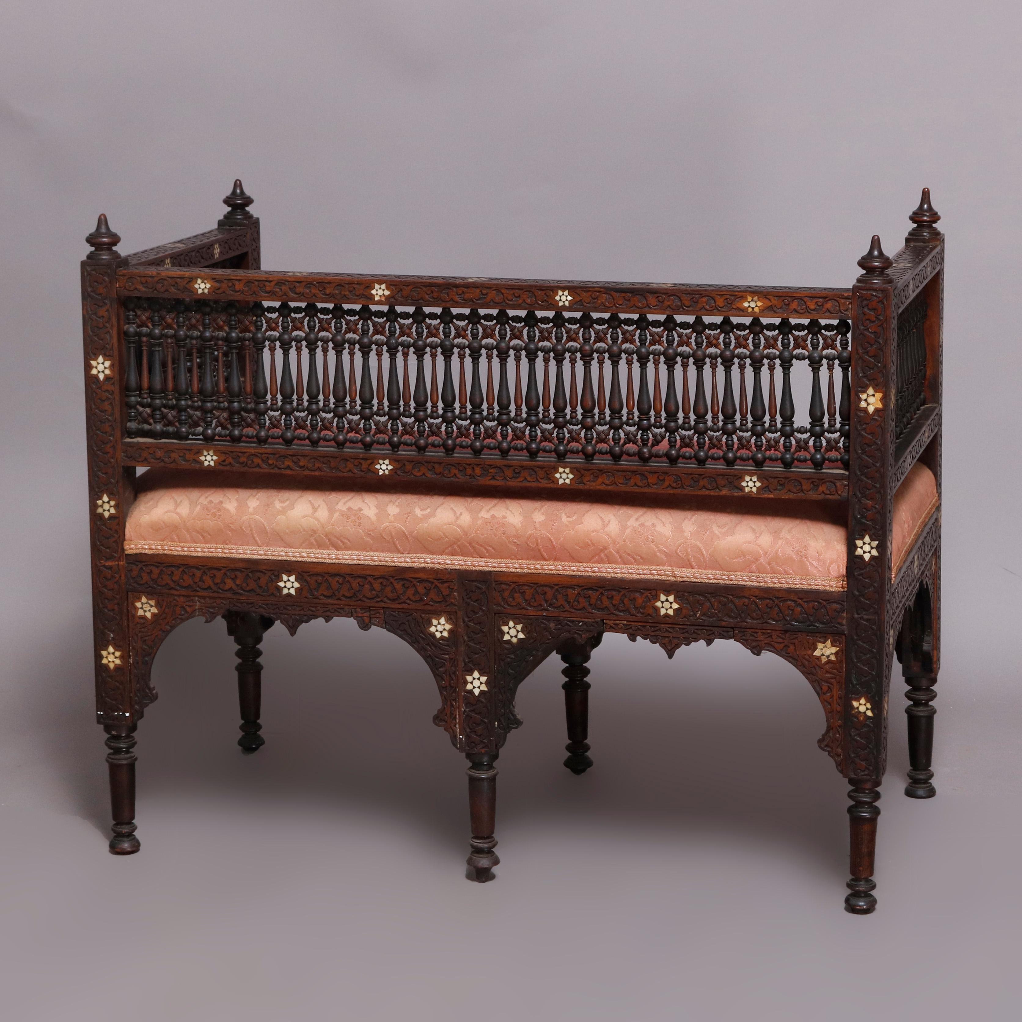 An antique Syrian Orientalist settee offers carved hardwood construction with back and arms having alternating shaped spindles with mother of pearl inlaid frame, upholstered seat and raised on carved and turned legs with arch form corbels, 19th