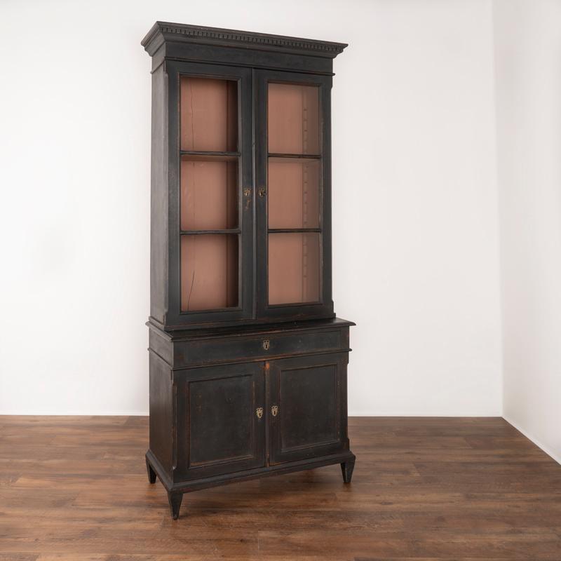 This antique black painted display cabinet from Denmark is made in 2 parts. The upper section has 2 adjustable shelves behind 2 glass doors and the bottom has one long narrow drawer and lower cabinet with one shelf. The newer applied black matte