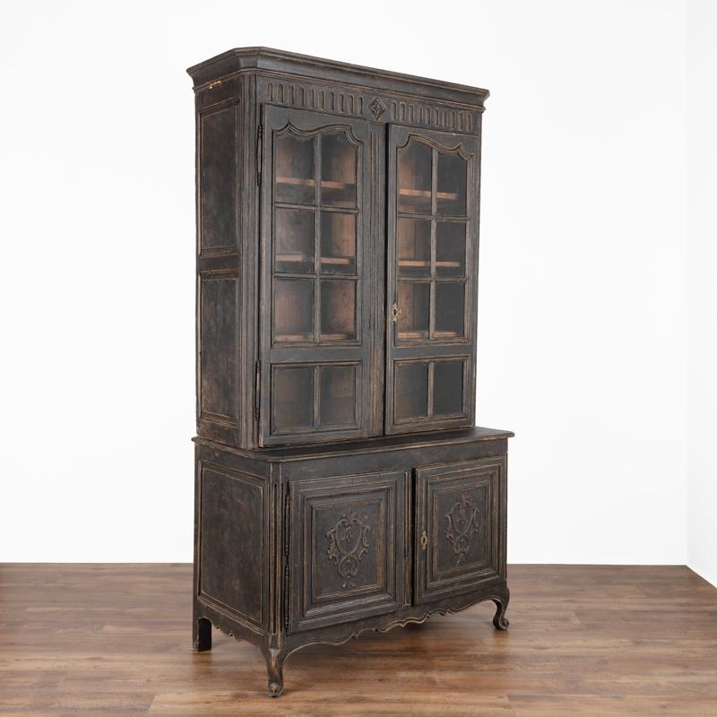 This striking antique oak cabinet was given a (newer) black painted finish, that has been distressed to compliment the age and French country style of the bookcase  and compliments the decorative carvings. The upper glass doors allow for beautiful
