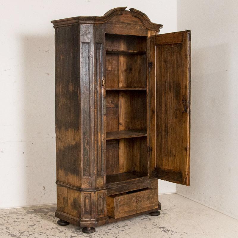 While primitive, there is a stately allure to this 7' tall single door armoire. The original paint has greatly been worn off through the distress of many years, leaving a residual amount of black and dark green paint, blended with large areas