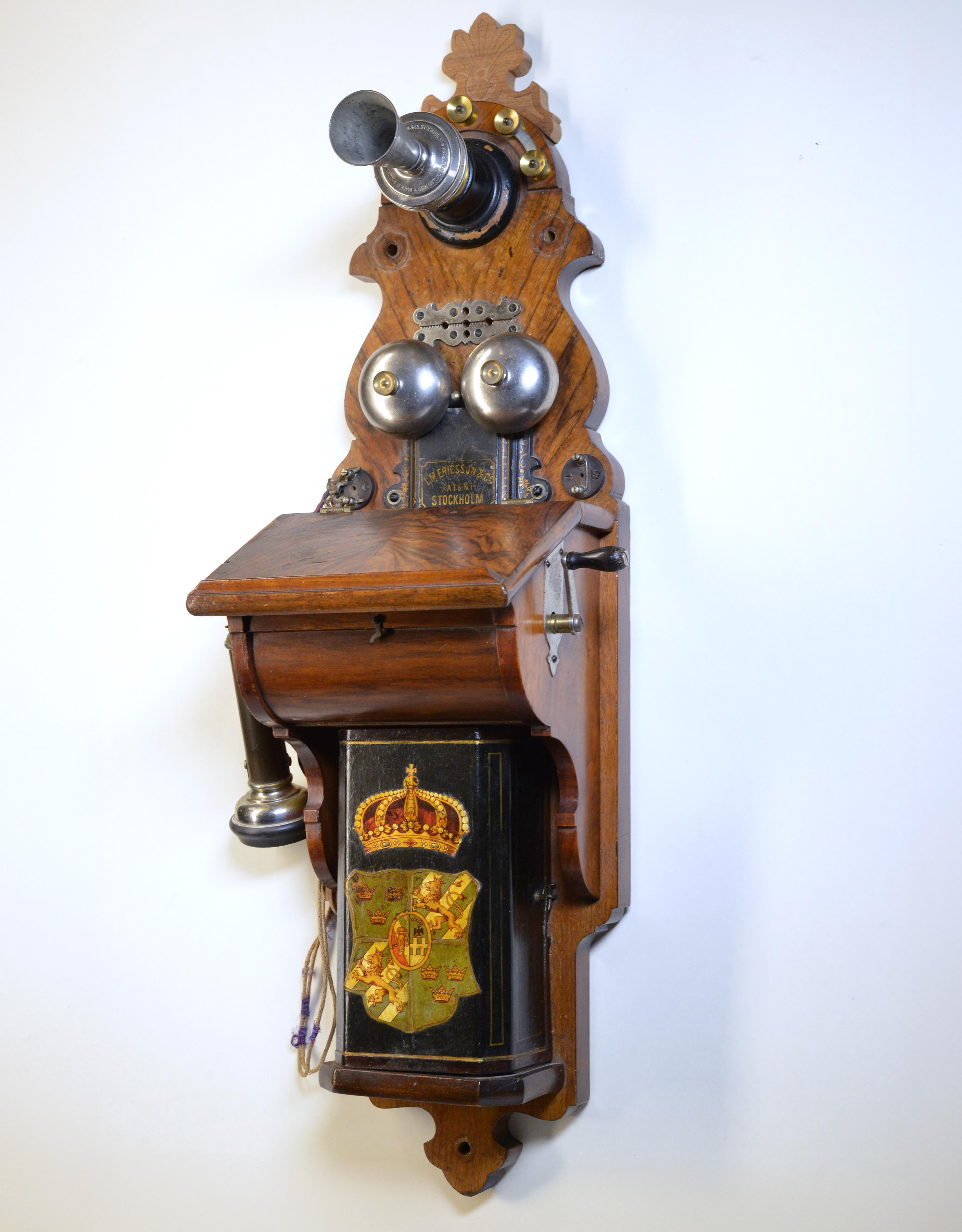 Telephone set, AB LM Ericsson, figure no. 1 or 3 in the 1889 catalogue, case in walnut, battery cover with lithographed decoration of large national coat of arms. Inside label stating that the phone was refurbished at the Royal Telegraph Office's