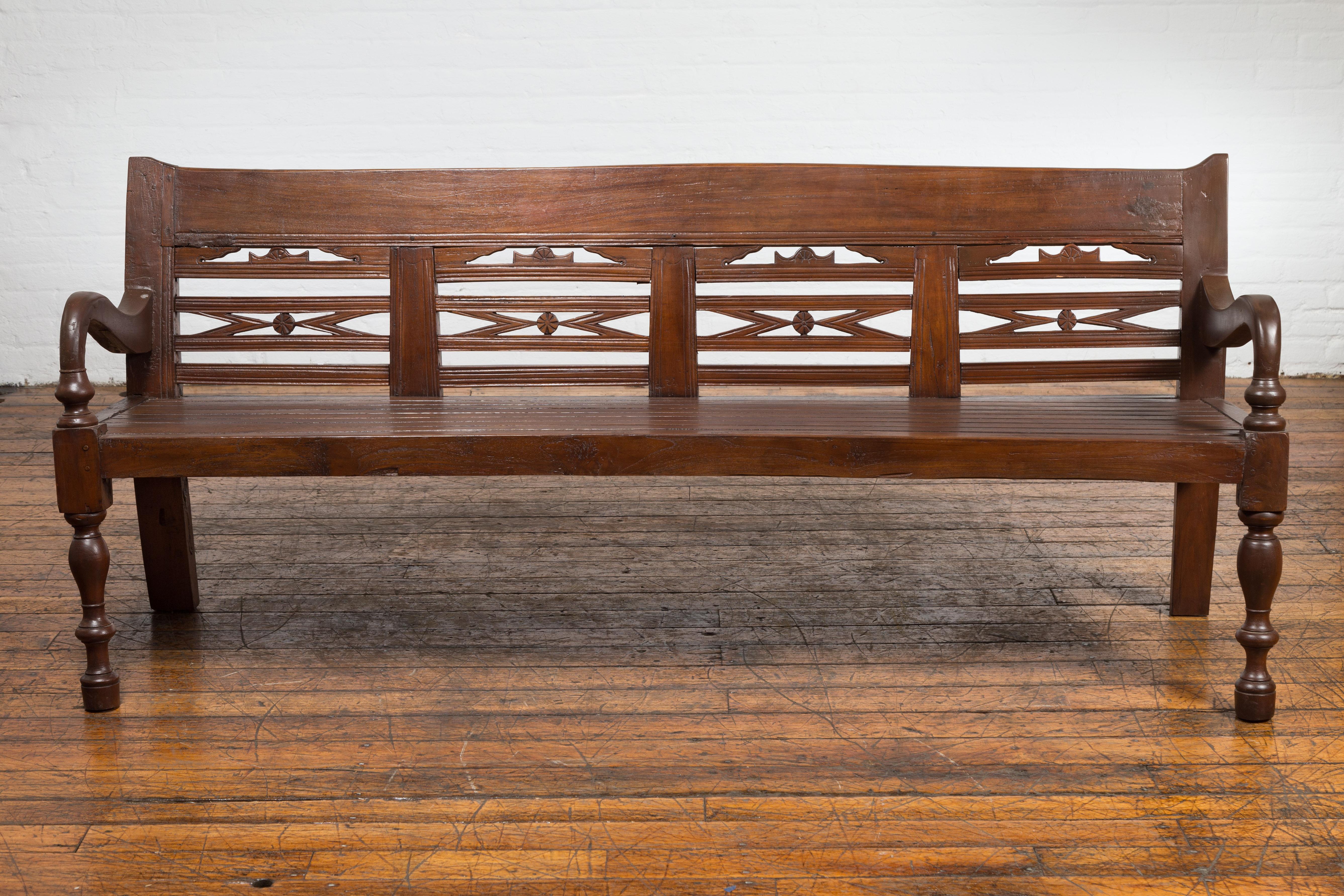 An antique teak wood Javanese settee from circa 1900 with hand-carved back, scrolling arms and baluster legs. This antique Javanese settee, dating back to circa 1900, is a striking piece of furniture crafted from teak wood, renowned for its