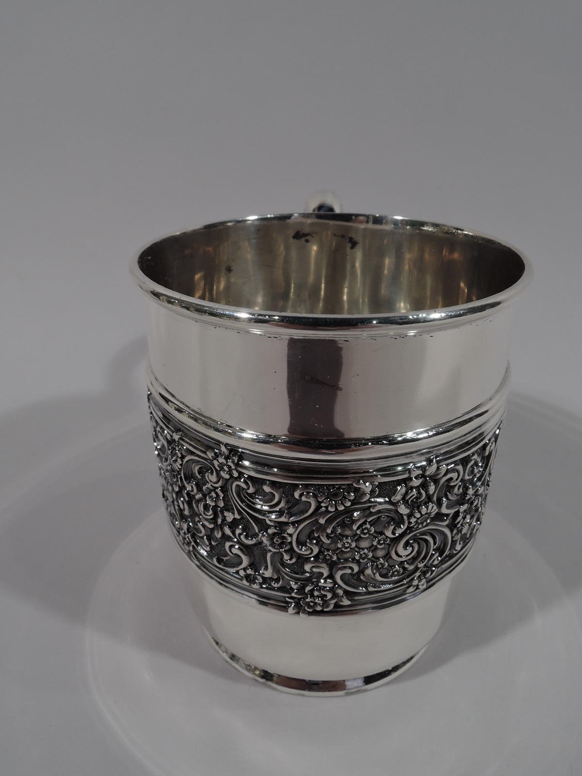 Edwardian sterling silver baby cup. Made by Tiffany & Co. in New York. Straight sides with wide ornamental band of scrolls, diaper, and flowers overlapping molded borders. High looping tapering handle with serrated leaf cap and short foot. Top and
