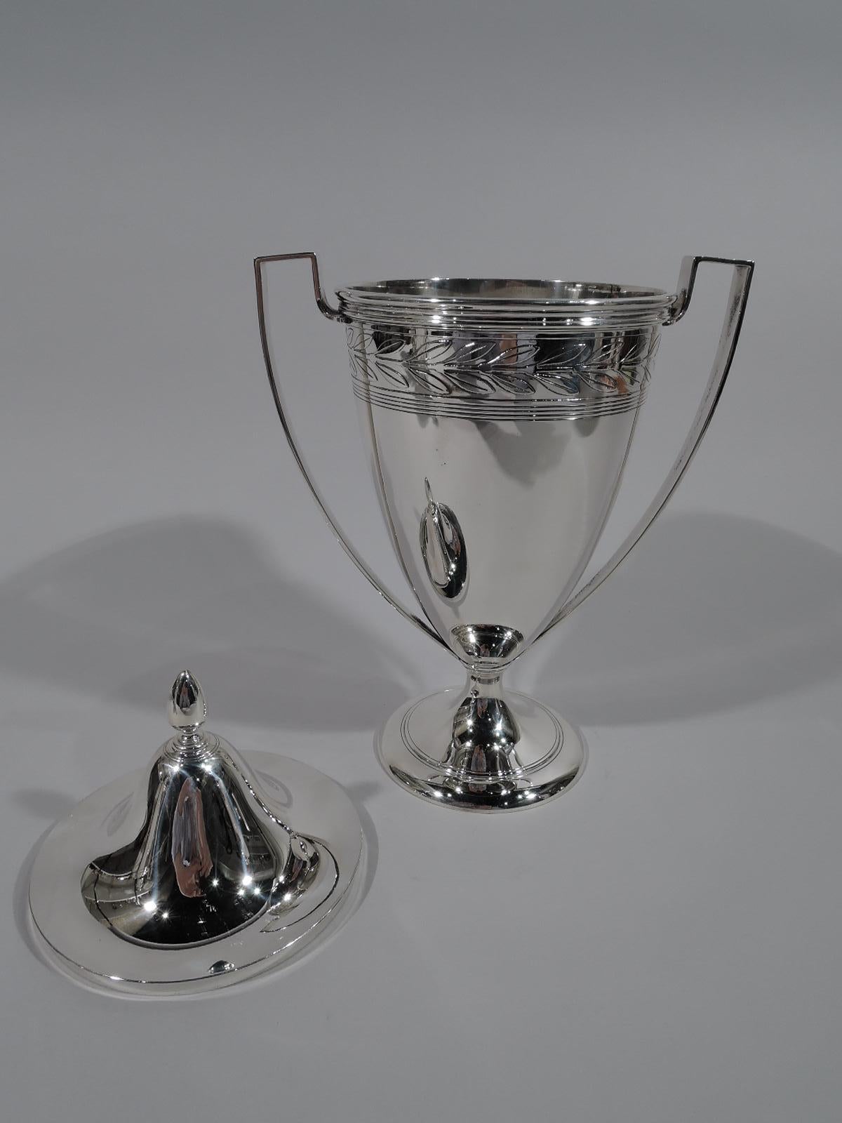 Art Deco sterling silver trophy cup. Made by Tiffany & Co. in New York, ca 1913. Ovoid with high-looping scroll bracket handles and raised foot. Cover domed with ovoid finial. Cup has acid-etched stylized laurel-wreath border at rim. Below there’s