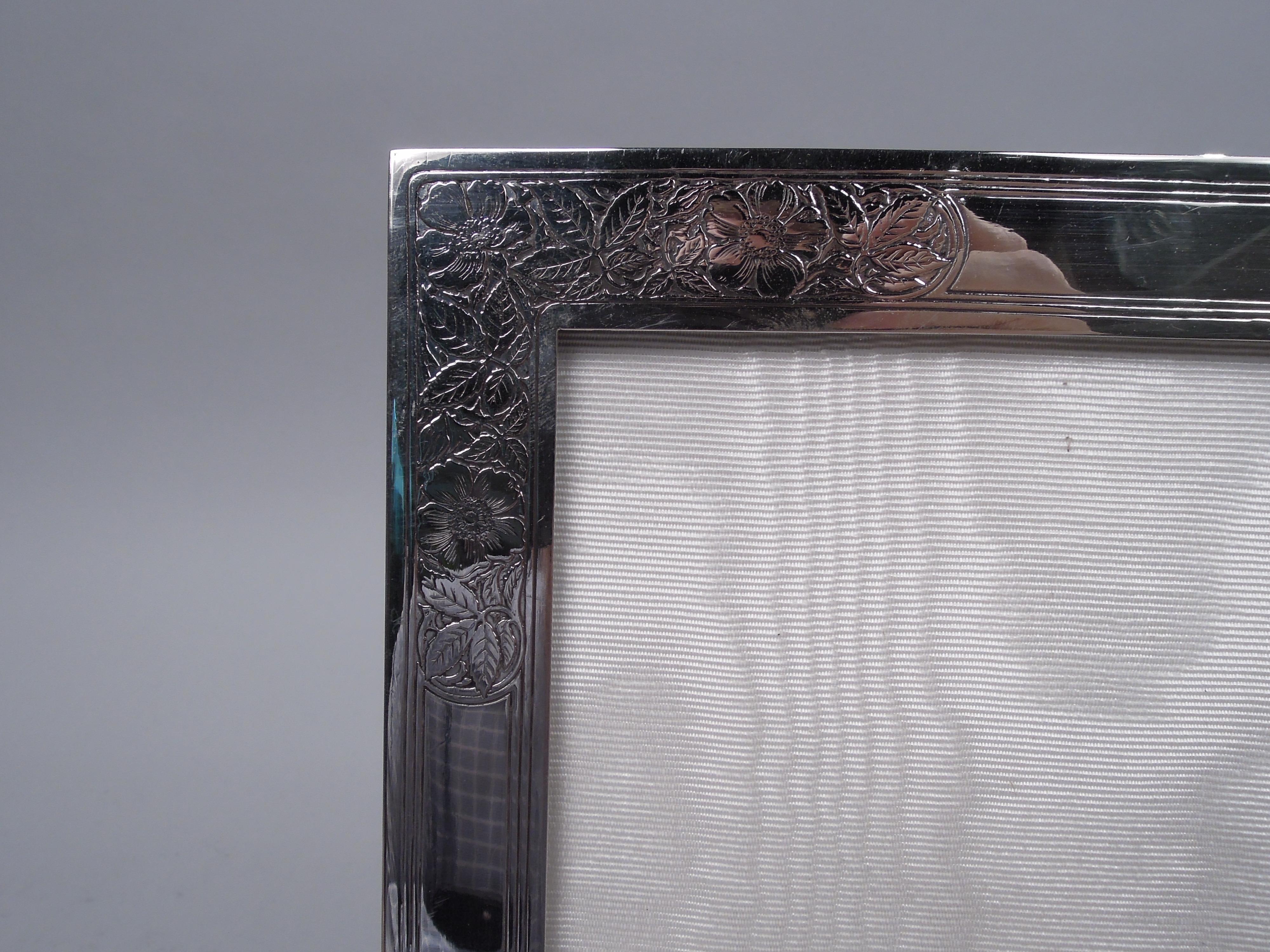 Art Nouveau sterling silver picture frame. Made by Tiffany & Co. in New York, ca 1910. Rectangular window in flat surround. Surround front has acid-etched fronds and flowers alternating with tubular cartouches (vacant). Sides have tooled rectilinear
