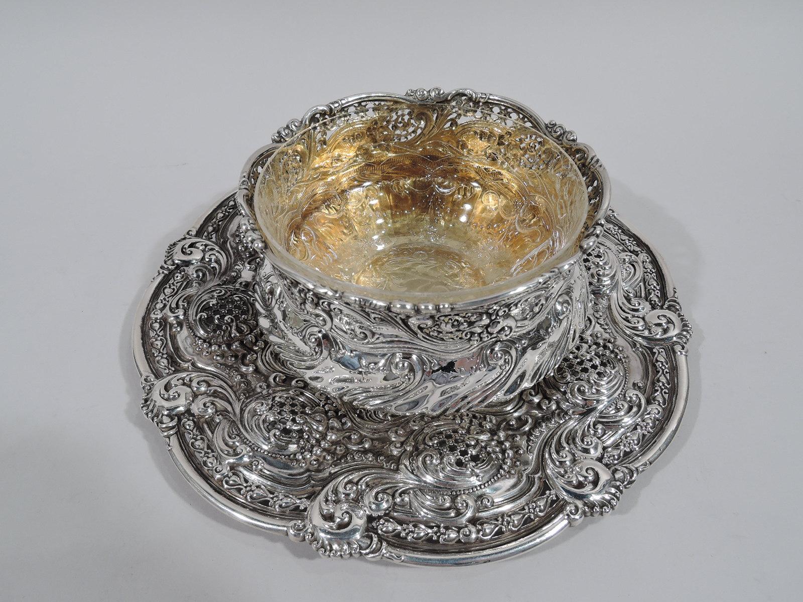 Sterling silver bowl on plate. Made by Tiffany & Co. in New York. Bowl: Squat with wavy and flared rim and short scrolled foot with bracket supports. Gilt interior. Plate: Round with scalloped rim. Plain and deep wavy star well with chased scrolls