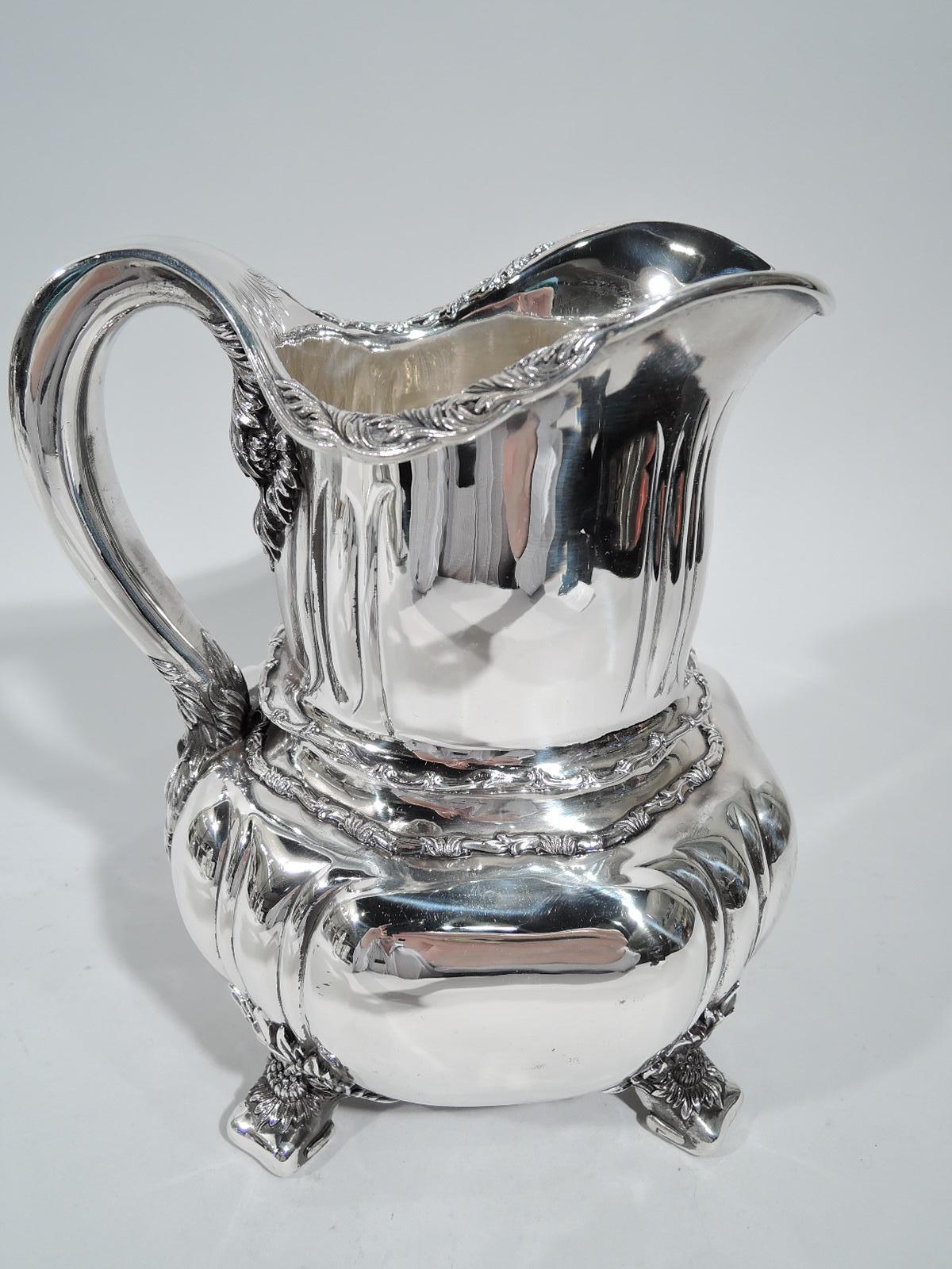 Chrysanthemum sterling silver water pitcher. Made by Tiffany & Co. in New York, ca 1910. Shaped bowl and curved neck with incised “slashes”. Helmet mouth and scroll handle. Rests on 4 flat volutes. Heavy and substantial with flower-head mounts on