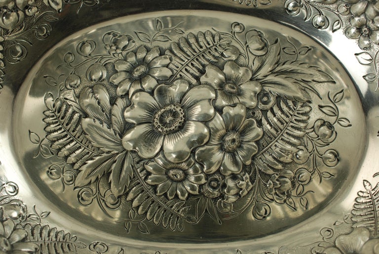 This elegant late 19th century sterling silver bowl was made by Tiffany & Company. The silver bowl has an oval shape and the interior features ornate repousse decoration in the form of ferns and flowers. The rim of the bowl has a scalloped wavy edge