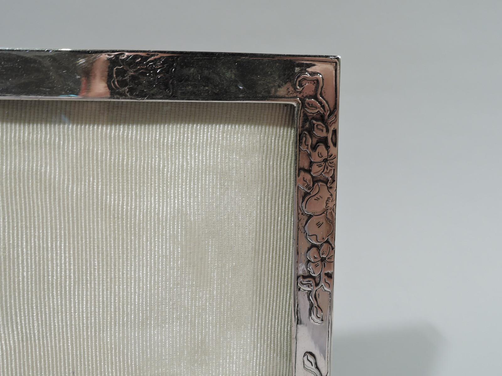 Edwardian Art Nouveau sterling silver picture frame. Made by Tiffany & Co in New York. Rectangular window in flat surround with acid-etched scattered blossoms and garlands on front and sides. At top central frame (vacant). With glass, silk lining,