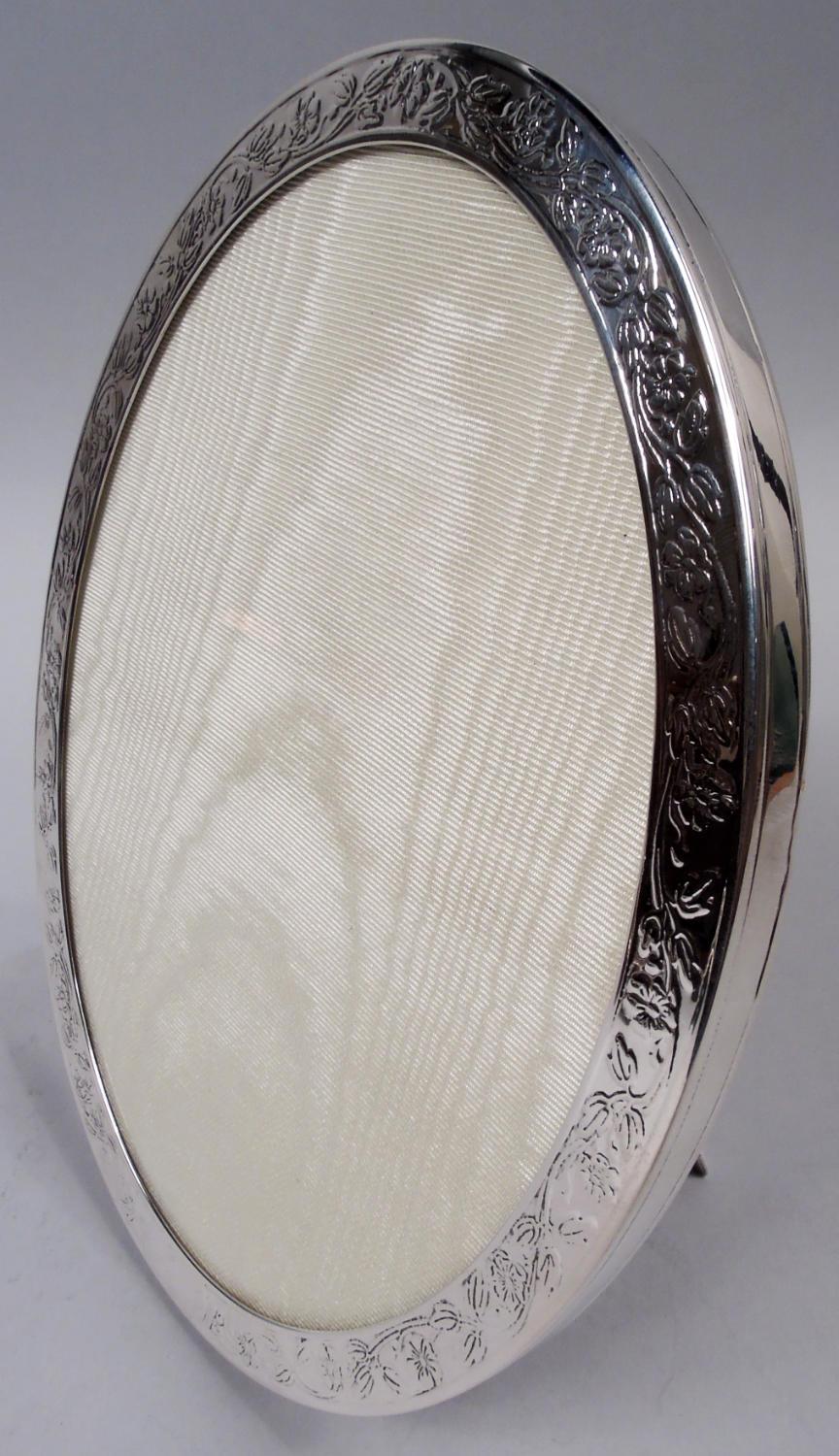 Edwardian sterling silver picture frame. Made by Tiffany & Co. in New York, ca 1910. Oval window in same surround; on front wraparound acid-etched floral garland; tubular cartouche (vacant). Sides plain with incised linear borders. With glass, silk