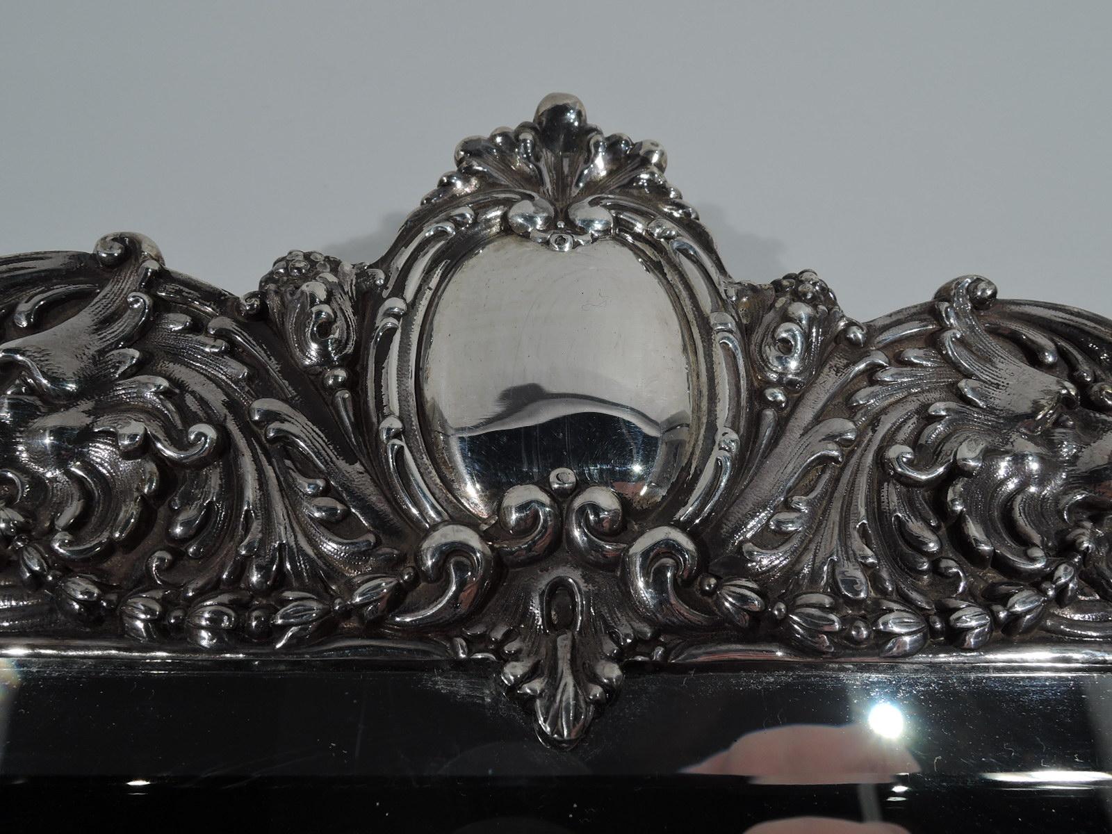 Rococo Revival sterling silver vanity mirror. Made by Tiffany & Co. in New York. Rectangular beveled glass in shaped surround with bracket supports and scrolled crown. Dense and dynamic ornament with scrolls and flowers. Crown has oval cartouche