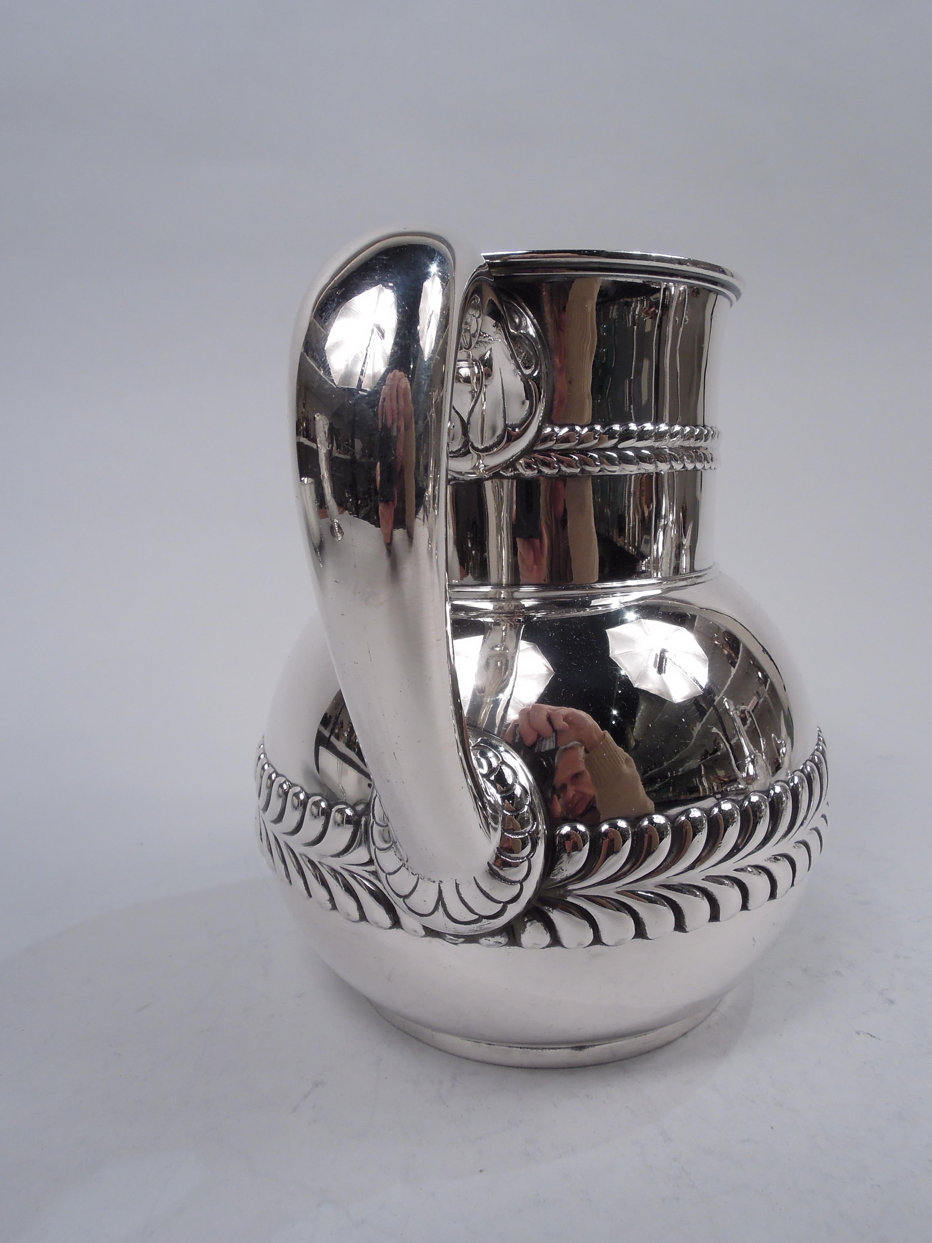 Victorian sterling silver water pitcher. Made by Tiffany & Co. in New York. Globular bowl and drum-form neck with small lip spout; c-scroll handle with stylized flower-head mounts. Bowl has embossed border of imbricated leaves or chevrons, a bold