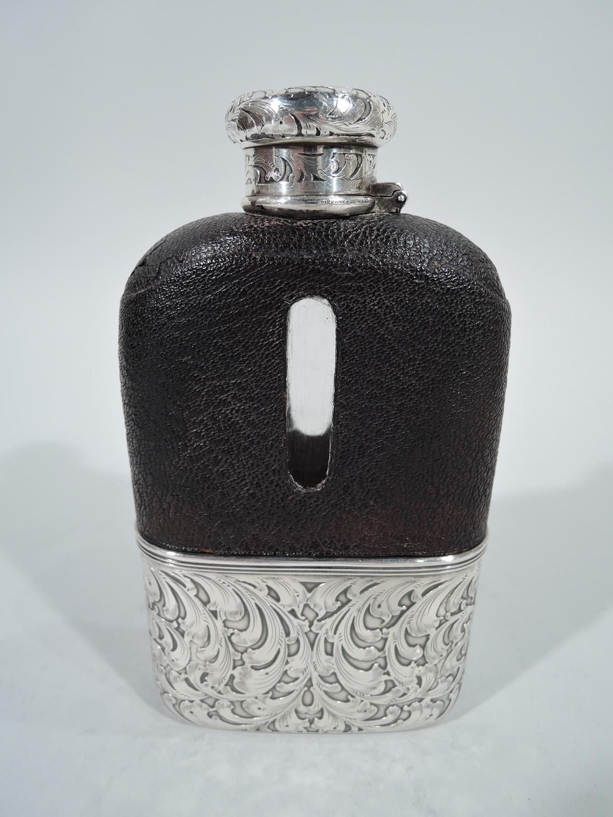 Edwardian safari flask. Made by Tiffany & Co. in New York. Rectilinear vessel in clear glass. Top half encased in leather with cut-out tubular windows. Bottom half encased in detachable sterling silver cup with acid-etched armorial lions and