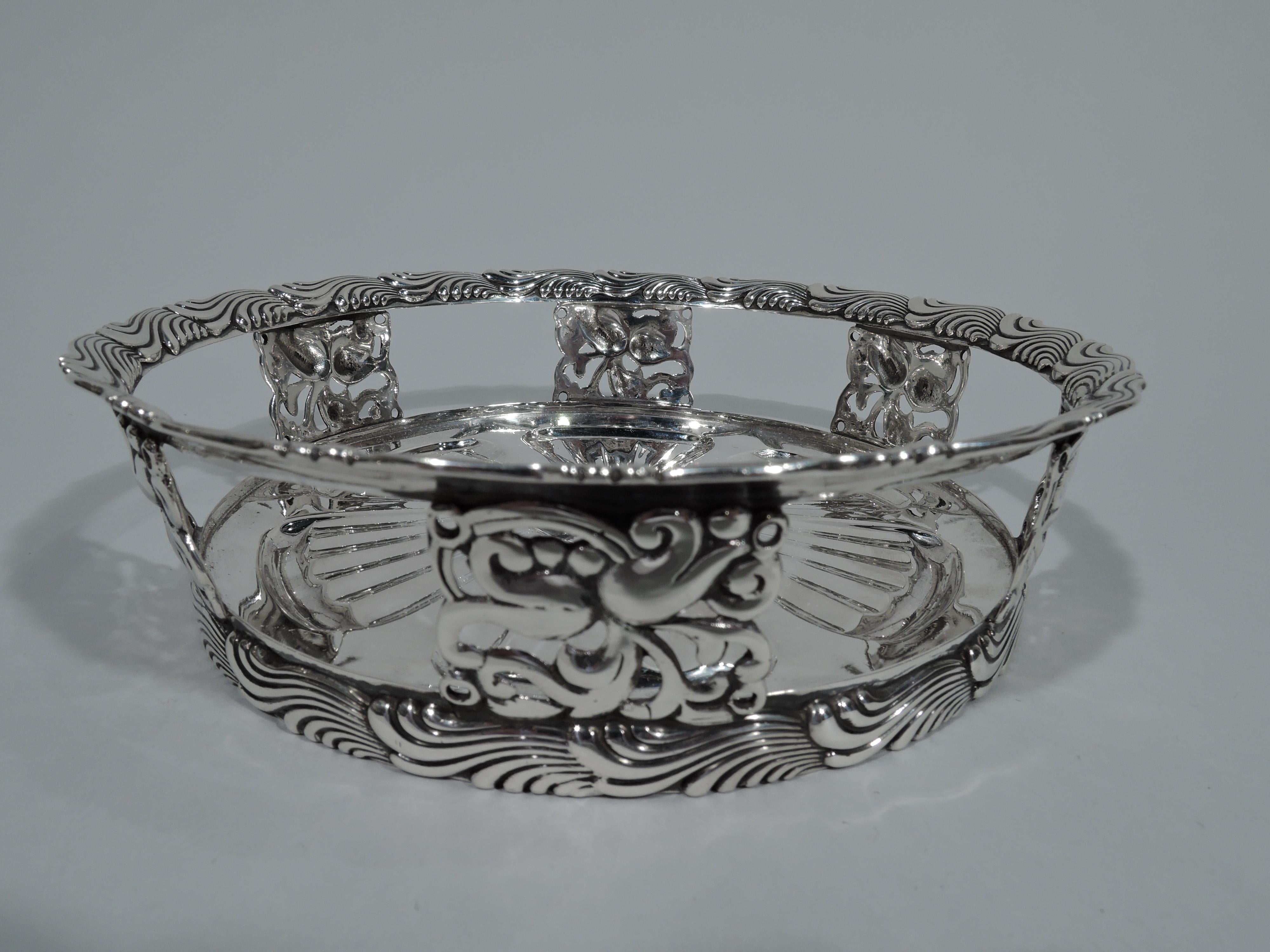 Stylish sterling silver wine bottle coaster. Made by Tiffany & Co. in New York. Solid well with fluted and radiating leaf and dart ornament. Open sides with blocks comprising pierced scrollwork and flowers. Flared rim with chased and fluid wavy