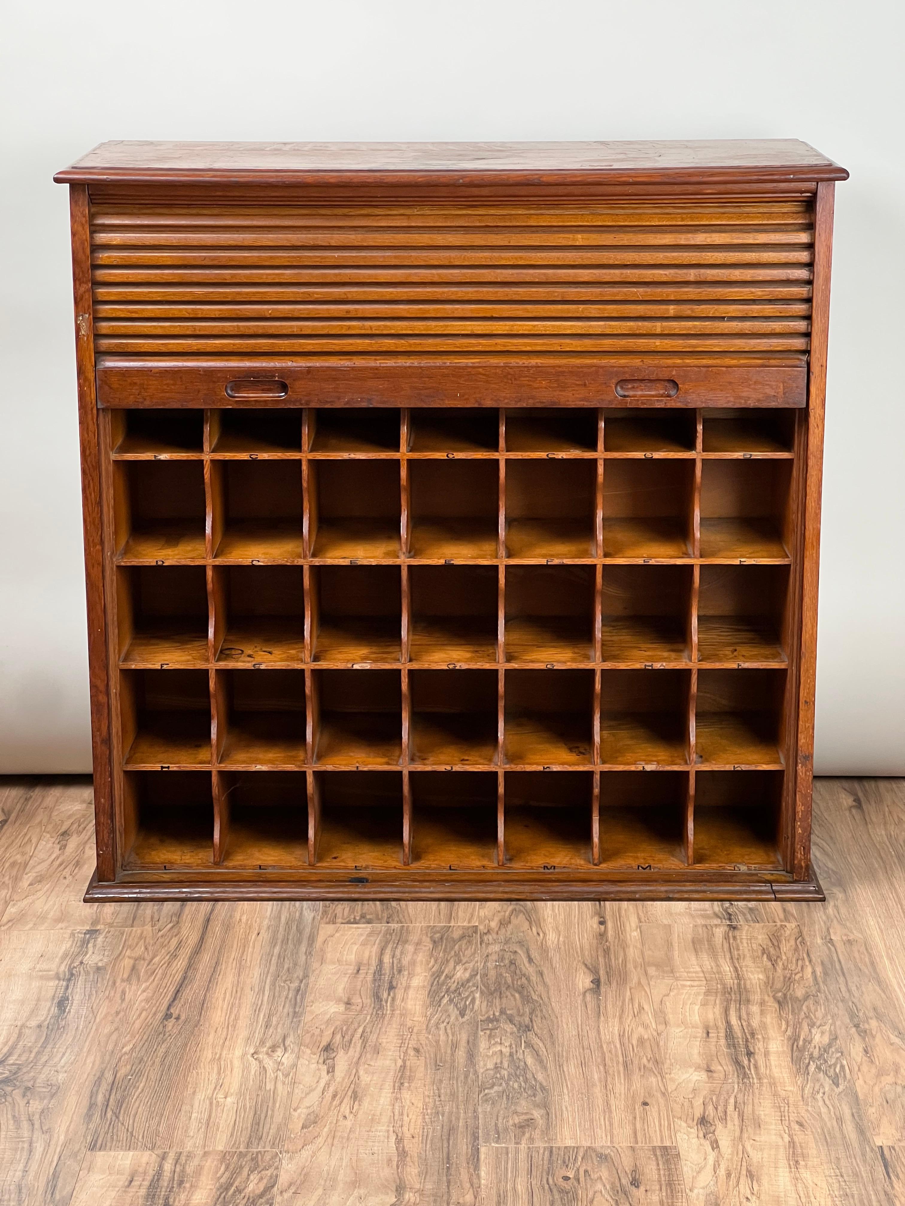 This Antique Tiger Oak Roll Top Pigeon Hole Cabinet, c. 1920, is a functional yet stylish piece that exemplifies the beauty of tiger oak wood, renowned for its bold grain and distinctive appearance. The cabinet features a unique roll-top design,