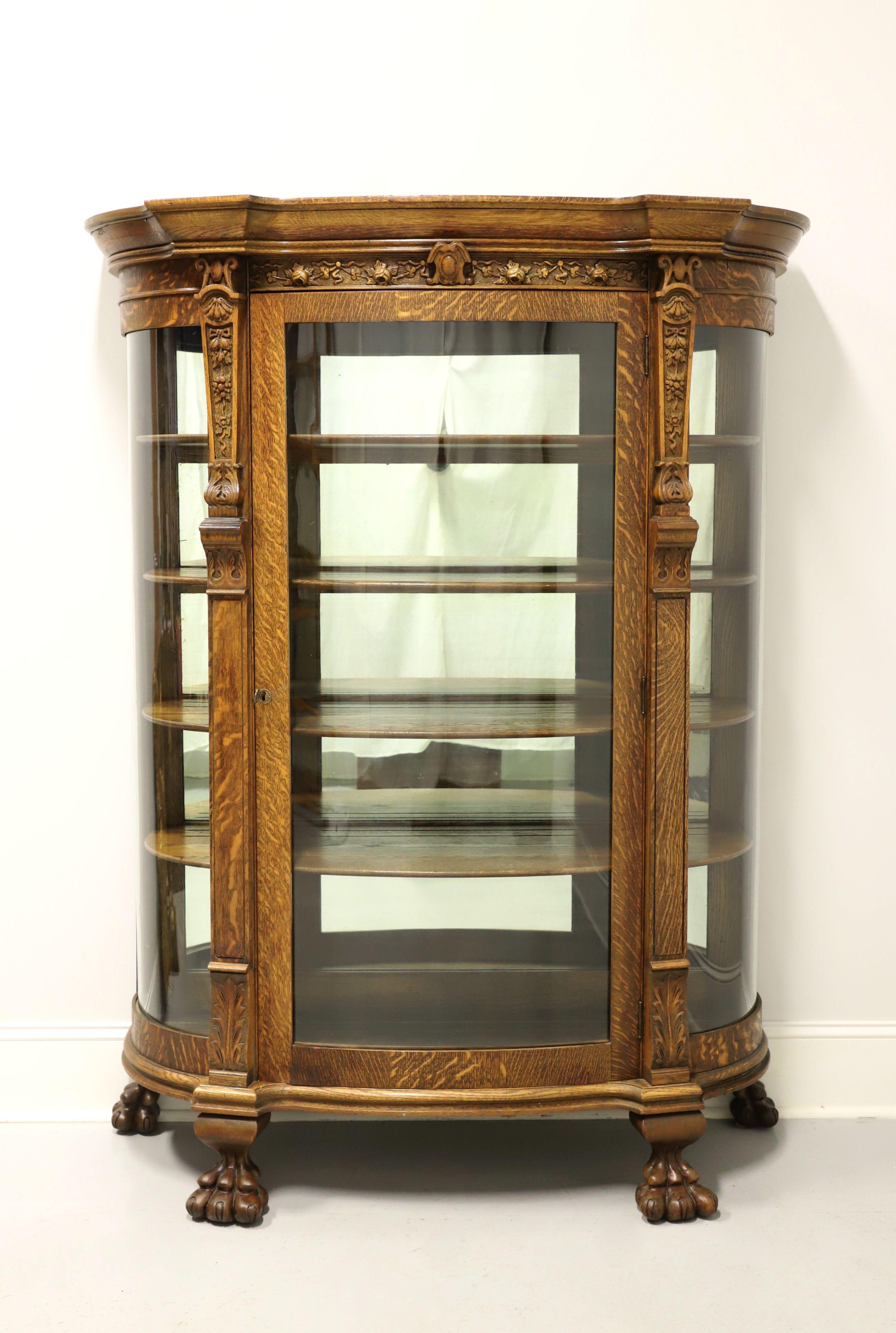 An antique bowfront china cabinet, unbranded. Tiger oak with decorative carvings, crown molding to top, carved columns flanking center door, brass keyhole escutcheon & key, and large paw feet. Cabinet features a single lockable door & side panels of