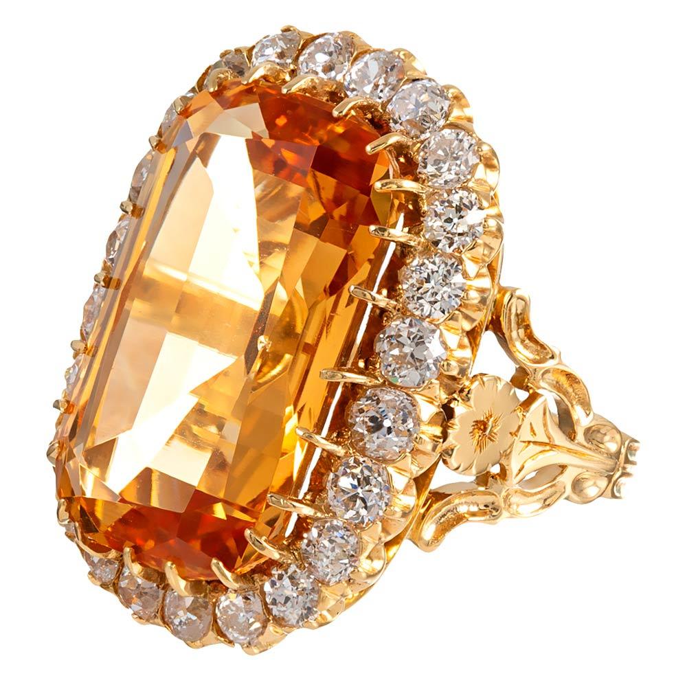 A 16 carat topaz exhibits exceptional gem-quality orange color from its antique golden setting, with white diamonds gleaming at its border. The major stone is accompanied by a GIA certificate. The diamonds weigh 2.40 carats in total and are ideally