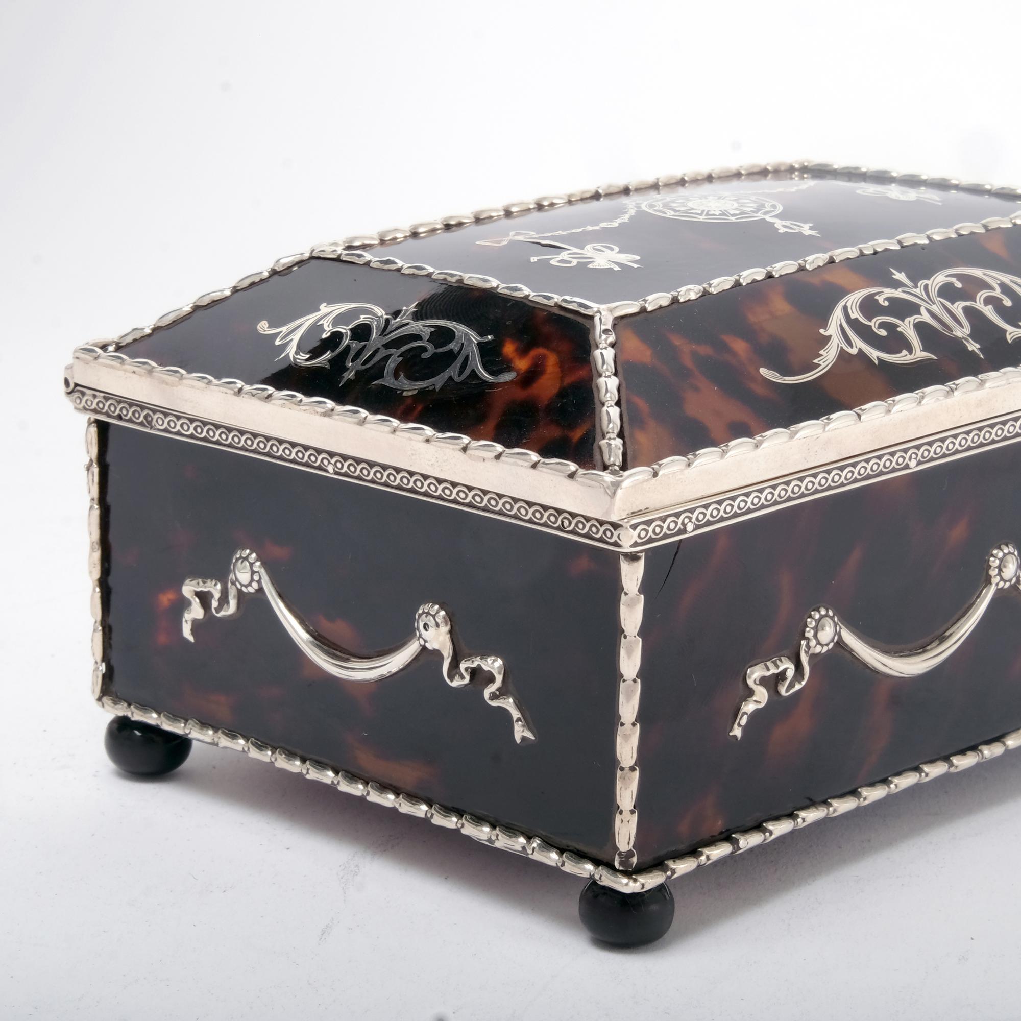 A fine Edwardian handmade jewel box with tortoiseshell panels held in place with a silver frame and decorated both with applied silver mounts in high relief plus silver inlay that has been hand engraved. The box rests on four ebonised wood bun