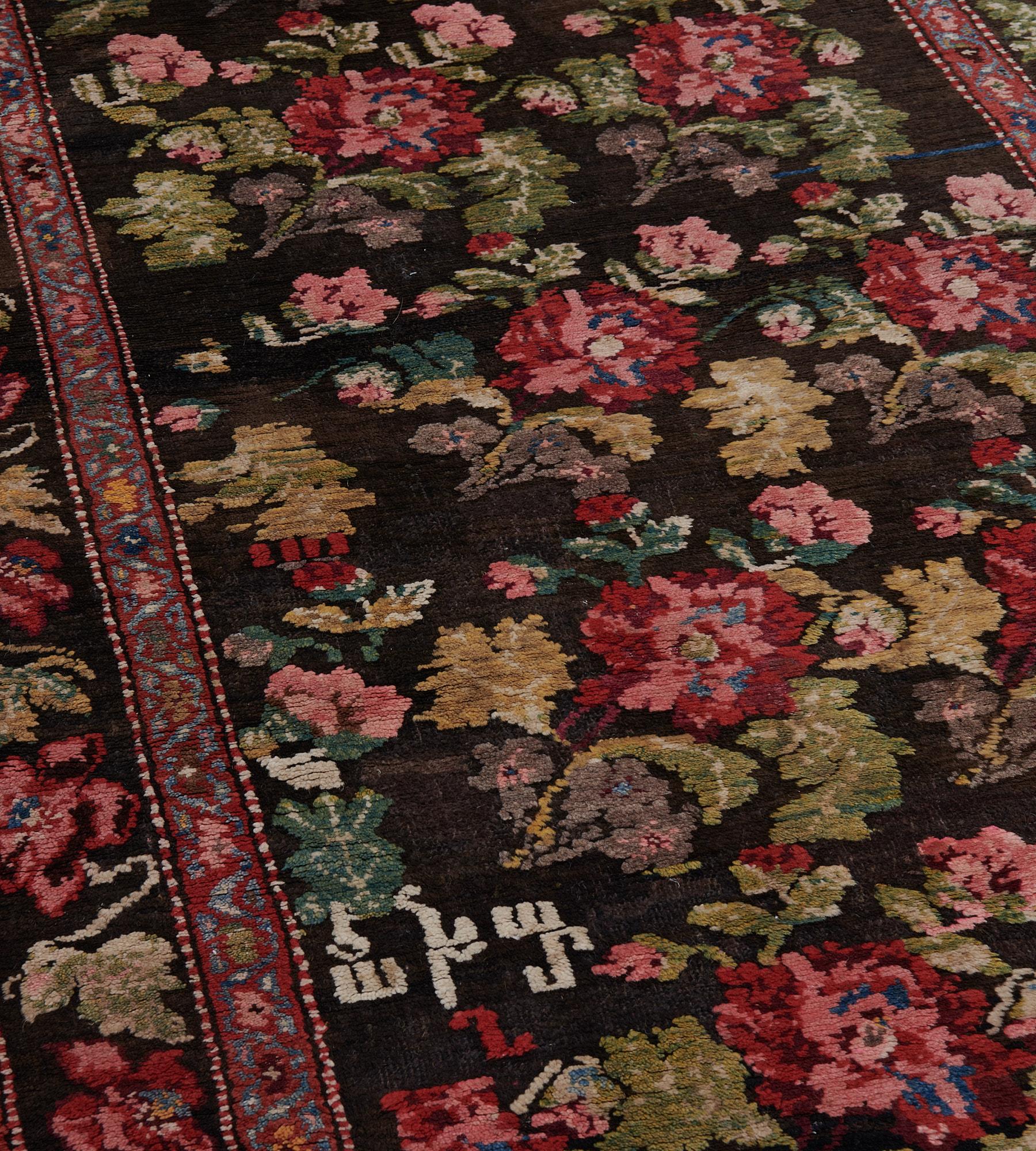 This antique Karabagh rug has a charcoal-black field with an overall design of cherry-red and dusty-pink floral bouquets with moss-green leaf and surrounded by similar smaller roses, an inscription band at one end and further letters to the lower