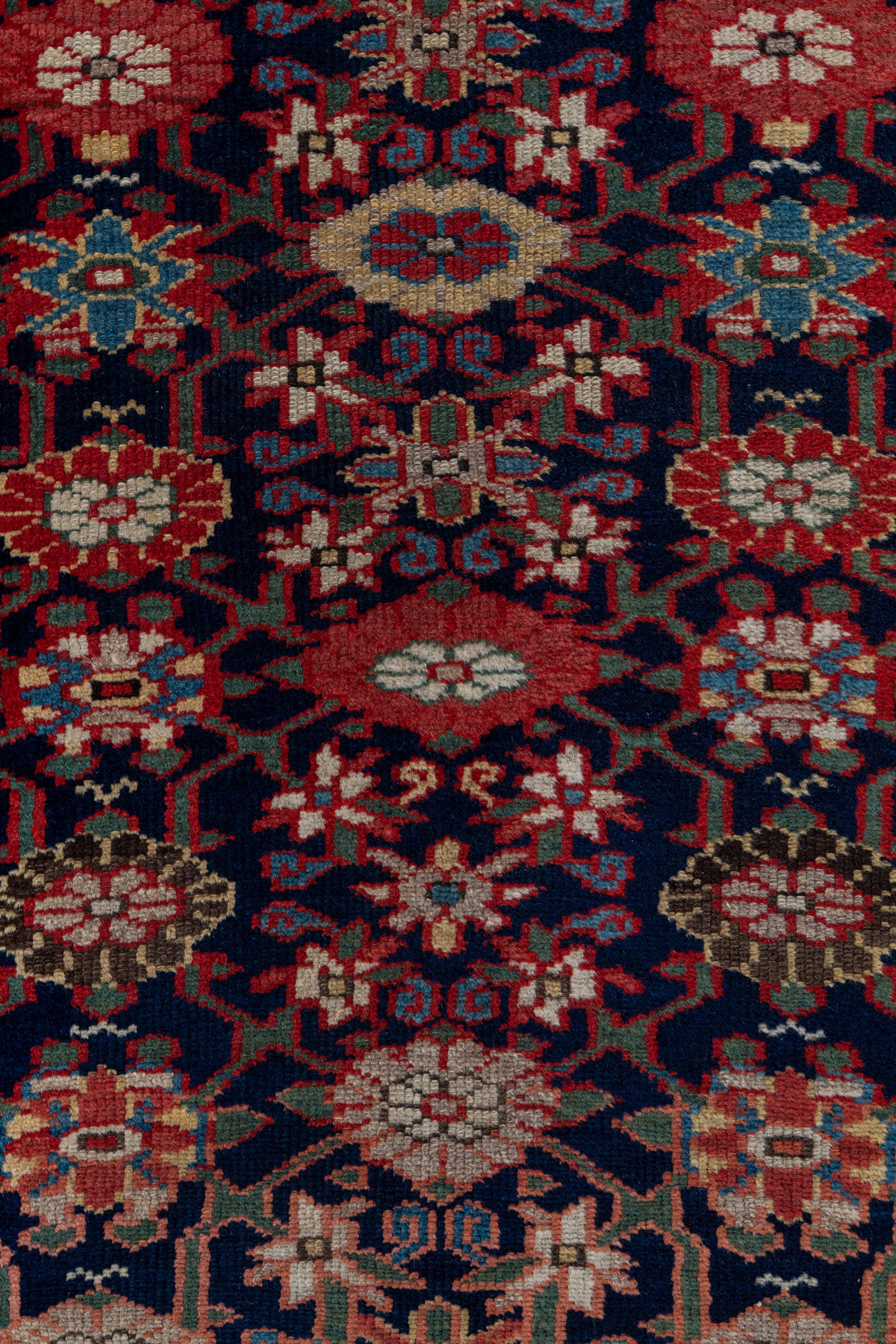 The navy field displays a slightly compressed version of the popular Mina Khani palmette lattice with polychrome larger elements and ecru small flowers. Rust border of reversing multicolor rosettes. Saturated palette. Medium rustic weave.