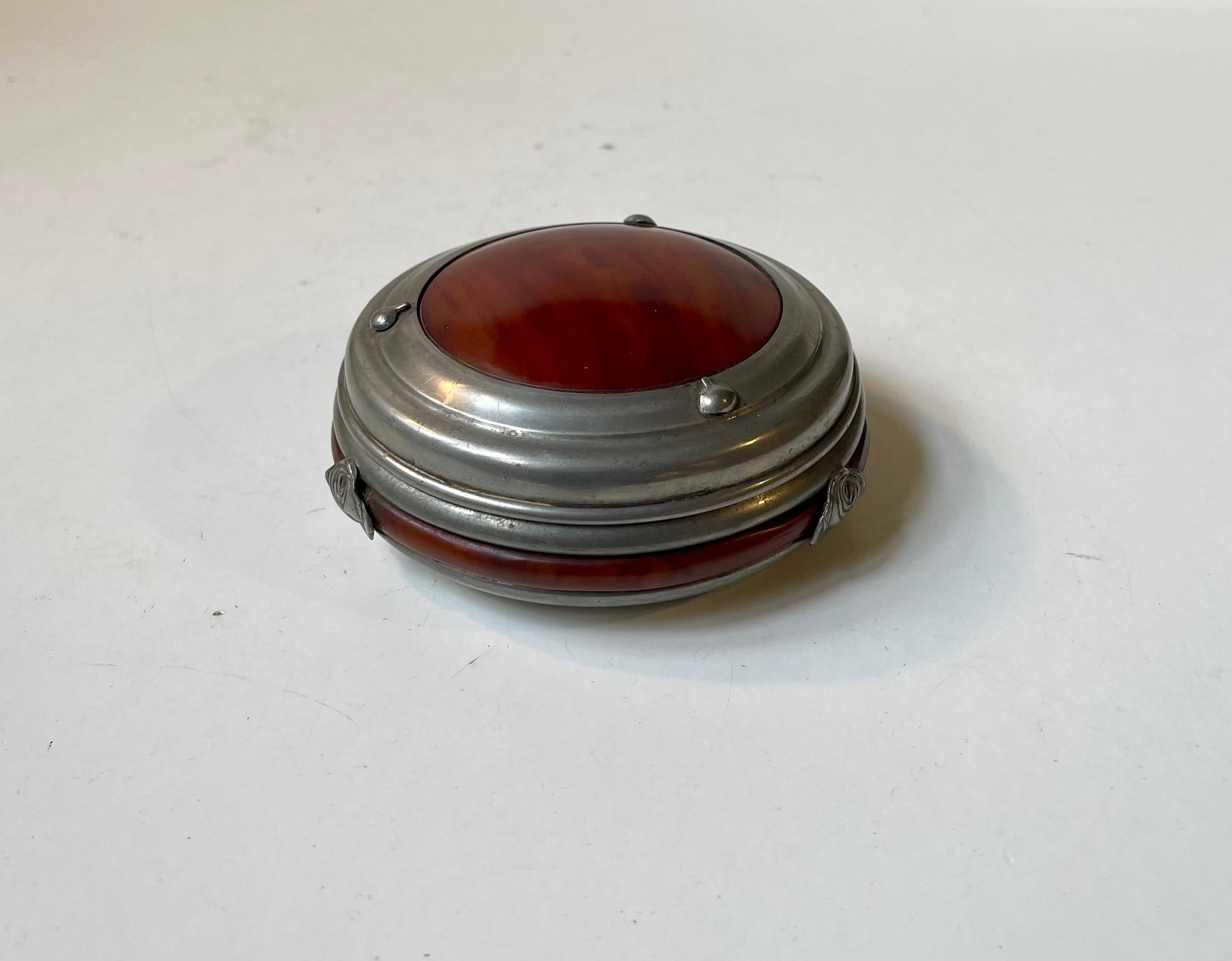 A stylish round container fashioned from pewter and inlaid with a red/brown material. Applied rose buds around its perimeter. Designed and made by Christian R. Kunst in Denmark circa 1900-20s. Marked beneath its base. 

Measurements: Diameter: 10