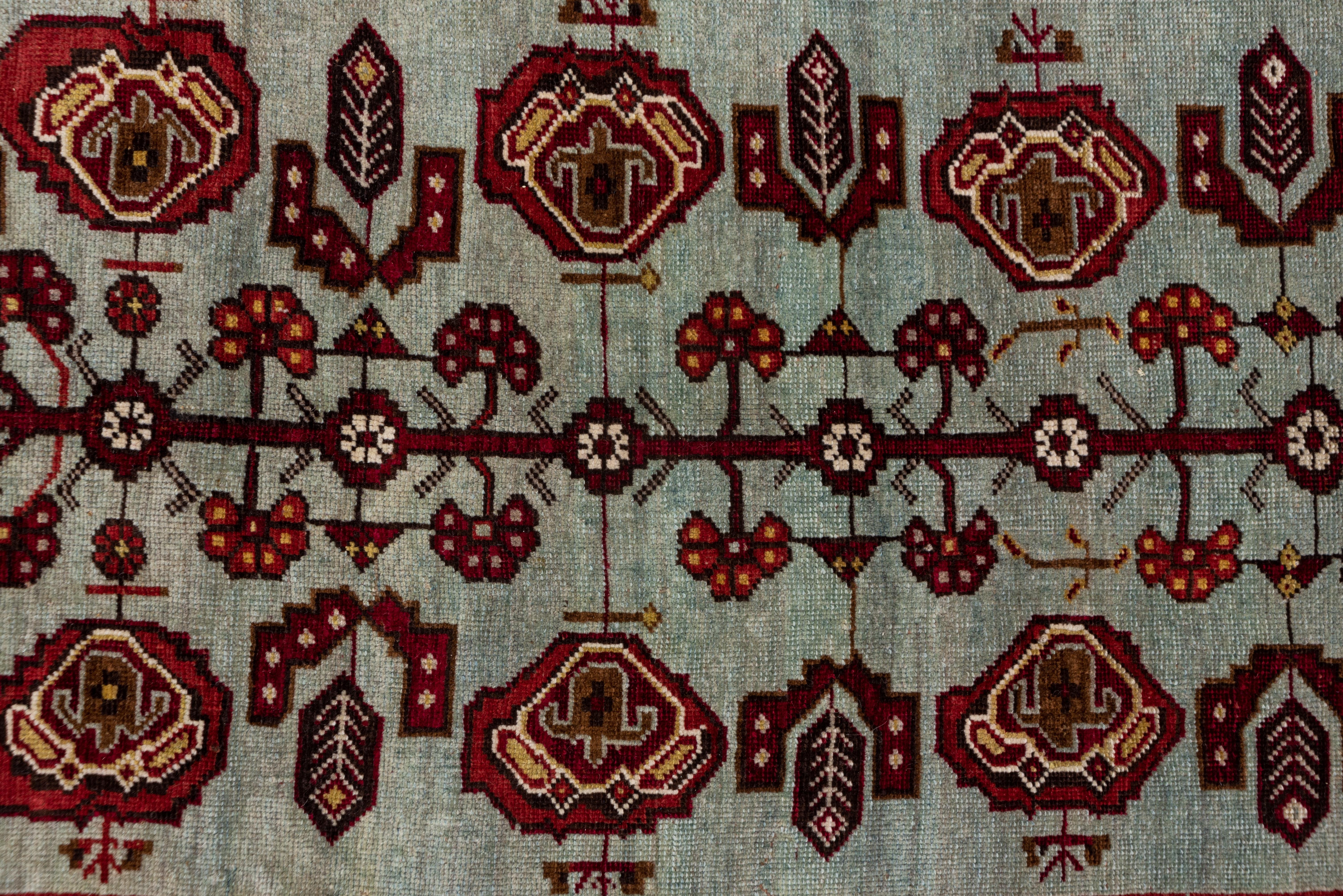 This Oushak rug has an intense wine-red field that displays a slender, geometric tendril flowing around the hexagonal pale blue-grey subfield with a central staff featuring carnations, palmettes, rosettes and leaves. The nicely abrashed green border