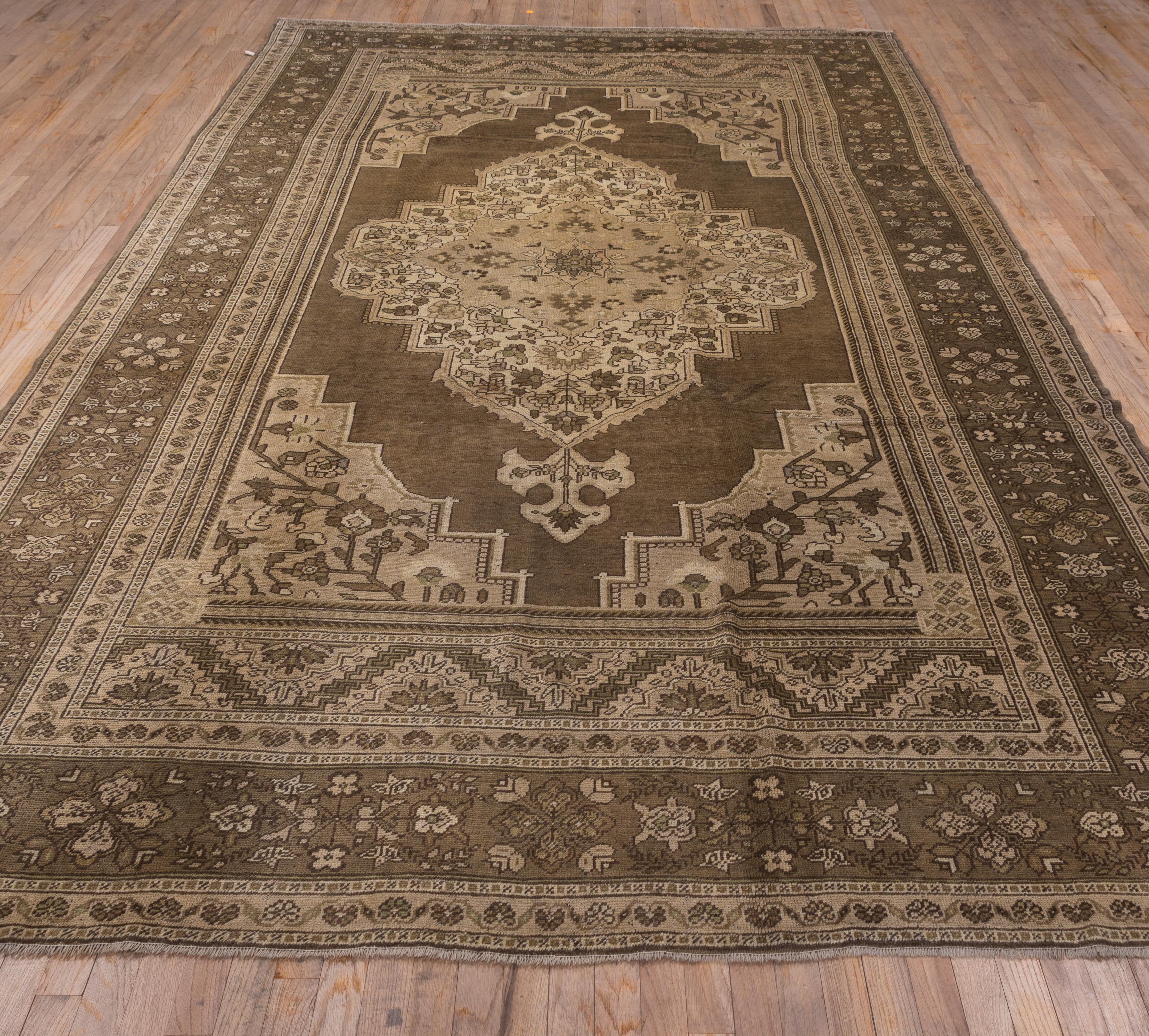 This moderately woven Turkish urban carpet shows extra zig-zag patterned panels at the field ends. The neutral open field displays an ecru stepped floral medallion with trefoil pendants. Border with floral crosses. Moderate weave.