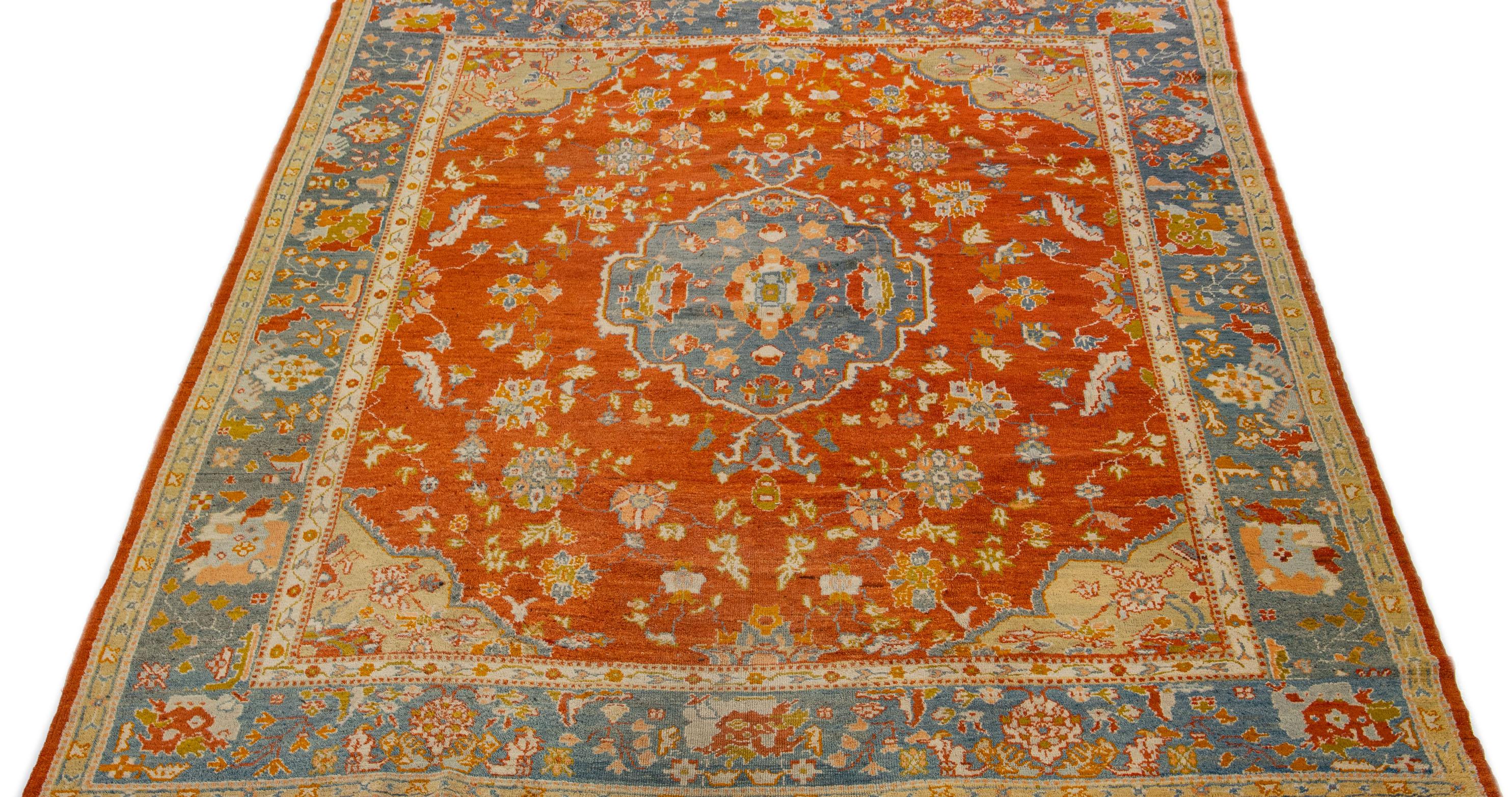 This Oushak wool rug boasts a stunning orange color field and is embellished with a navy blue frame and multicolored accents in a captivating medallion design. Its exquisite antique craftsmanship, and alluring aesthetics make it a delightful