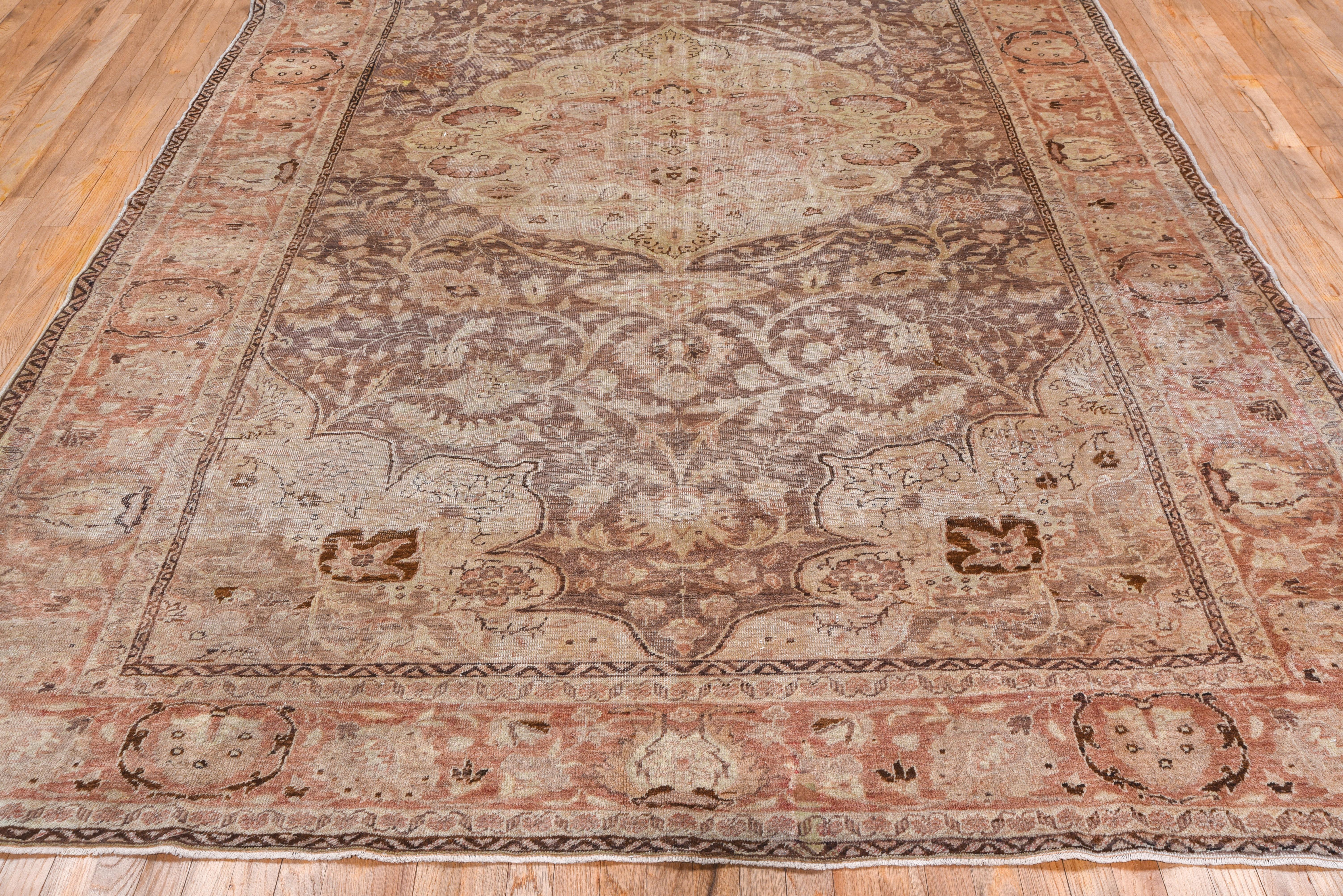 Wholly in the curvilinear Persian style, this moderately woven city carpet shows a large lightly scalloped and end-pointed ecru medallion, filled with palmettes and with doubled pendants, on a brown field with strong arabesques, ragged palmettes and