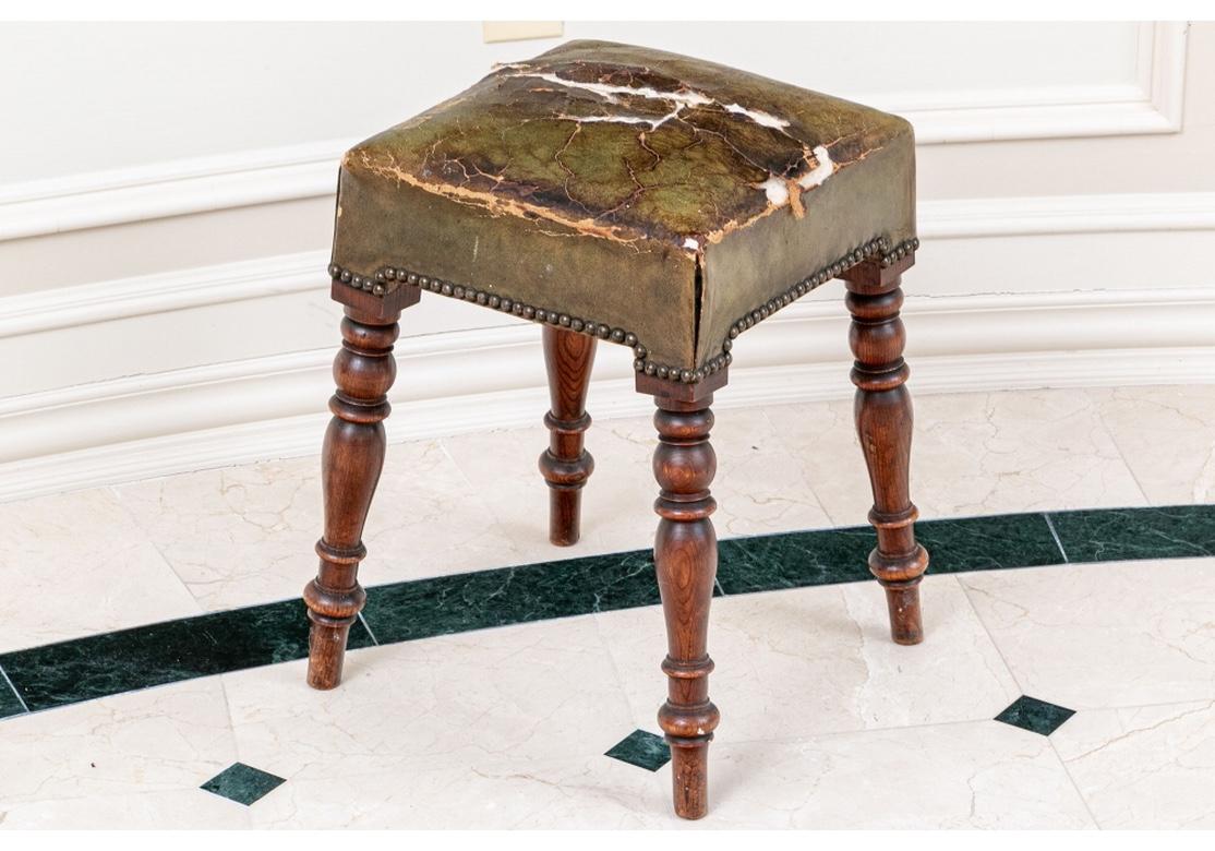 A carved and turned frame with slightly splayed legs raised on tall peg feet. The top upholstered in an olive green leather, now in need of replacement. Nail head trim. 
Measures: H. 19