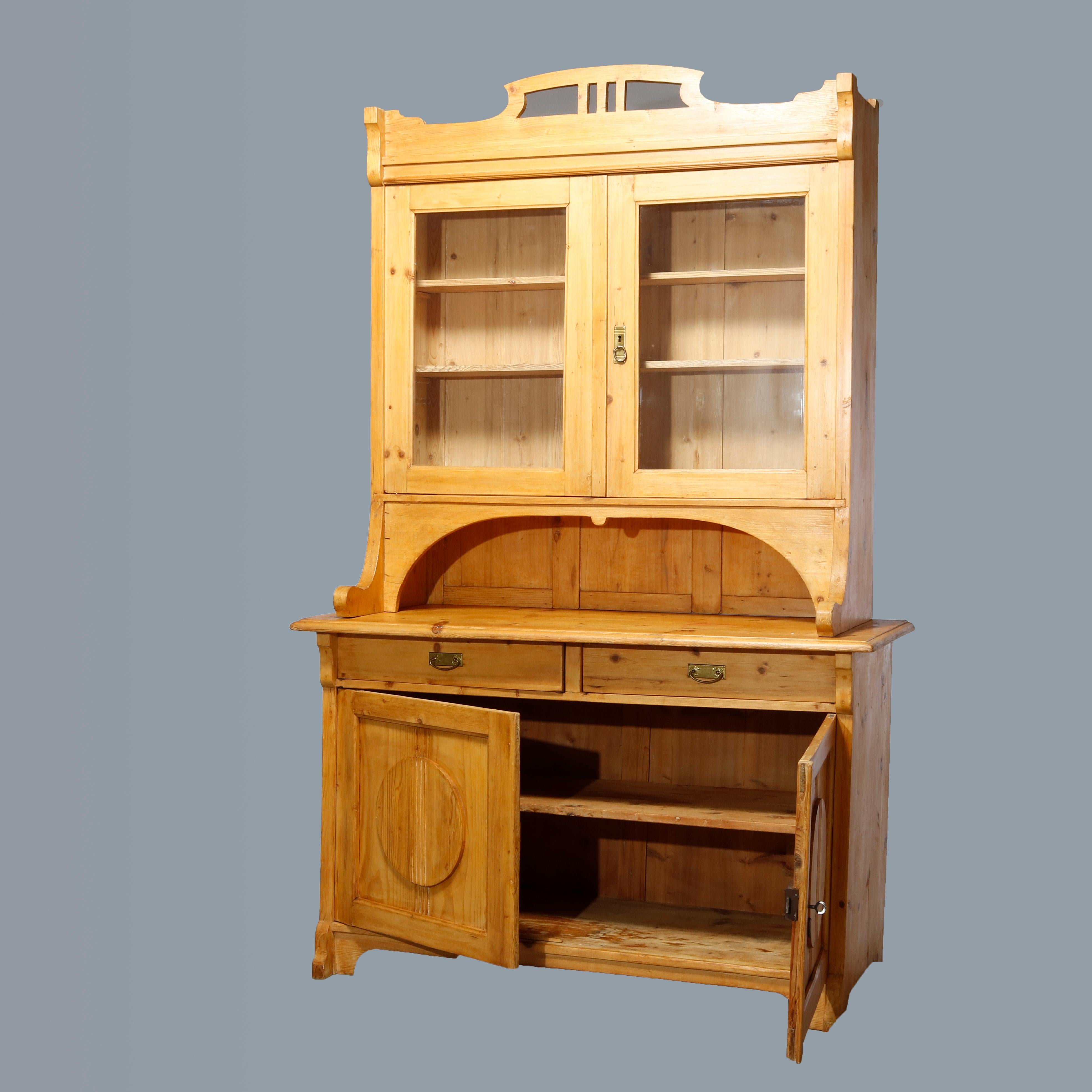 An antique step-back cupboard offers Austrian pine construction with upper hutch having pierced crest over double glass door cabinet surmounting lower cabinet having two drawers over cabinet having double paneled doors with circular reserves,
