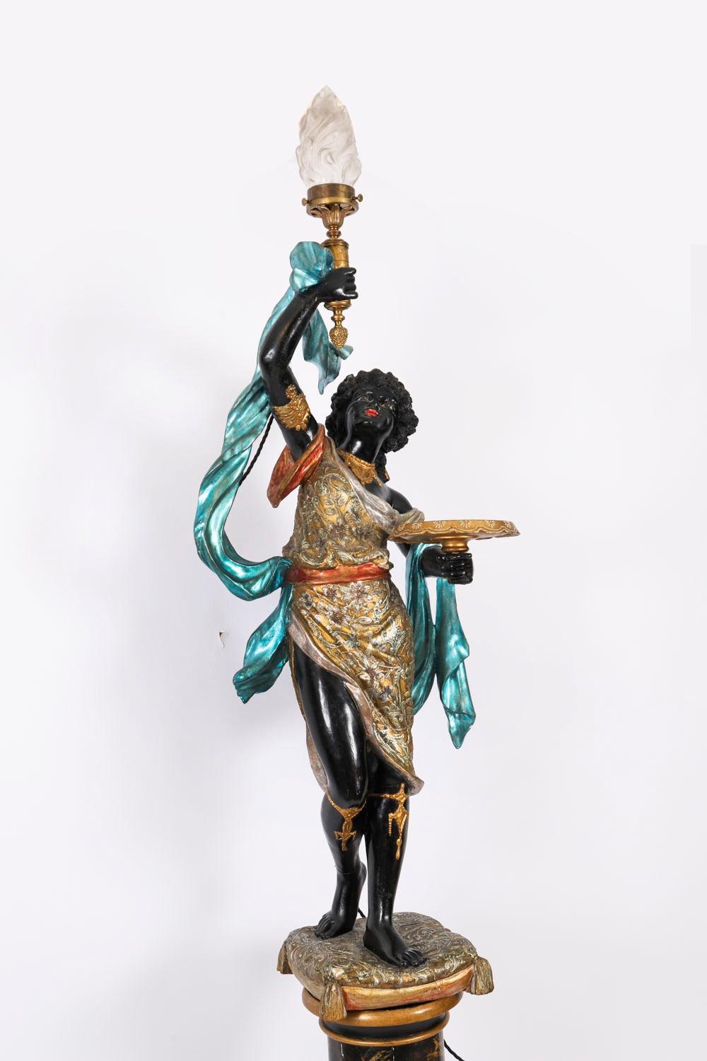 Se trata de una hermosa antorcha figurada veneciana antigua de madera tallada, dorada y policromada, sobre peana, de hacia 1880.

La torchere está modelada como una mujer de pie sobre un cojín ornamentado que sostiene un plato en una mano y una