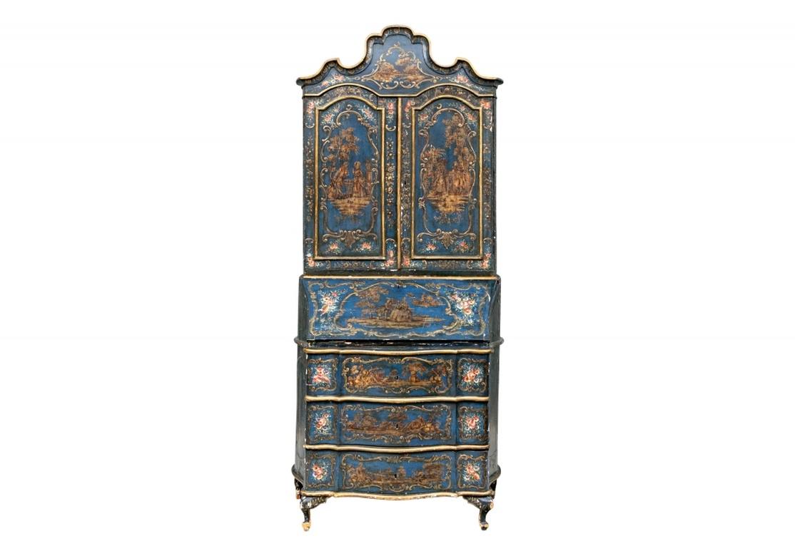 A tall cabinet made in two parts in blue paint with polychrome painted floral and gilt figural decoration. With a serpentine baroque crest with a gilt landscape in the center cartouche flanked by colorful flowers. The two door cabinet with gilt