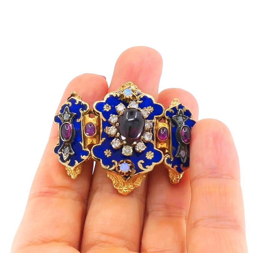 Un sorprendente gioiello antico a forma di fibbia realizzato in oro 14k, evidenziato da uno smalto blu intenso e incastonato da una straordinaria serie di pietre preziose. Al centro si trova un granato ovale cabochon, circondato da diamanti, mentre