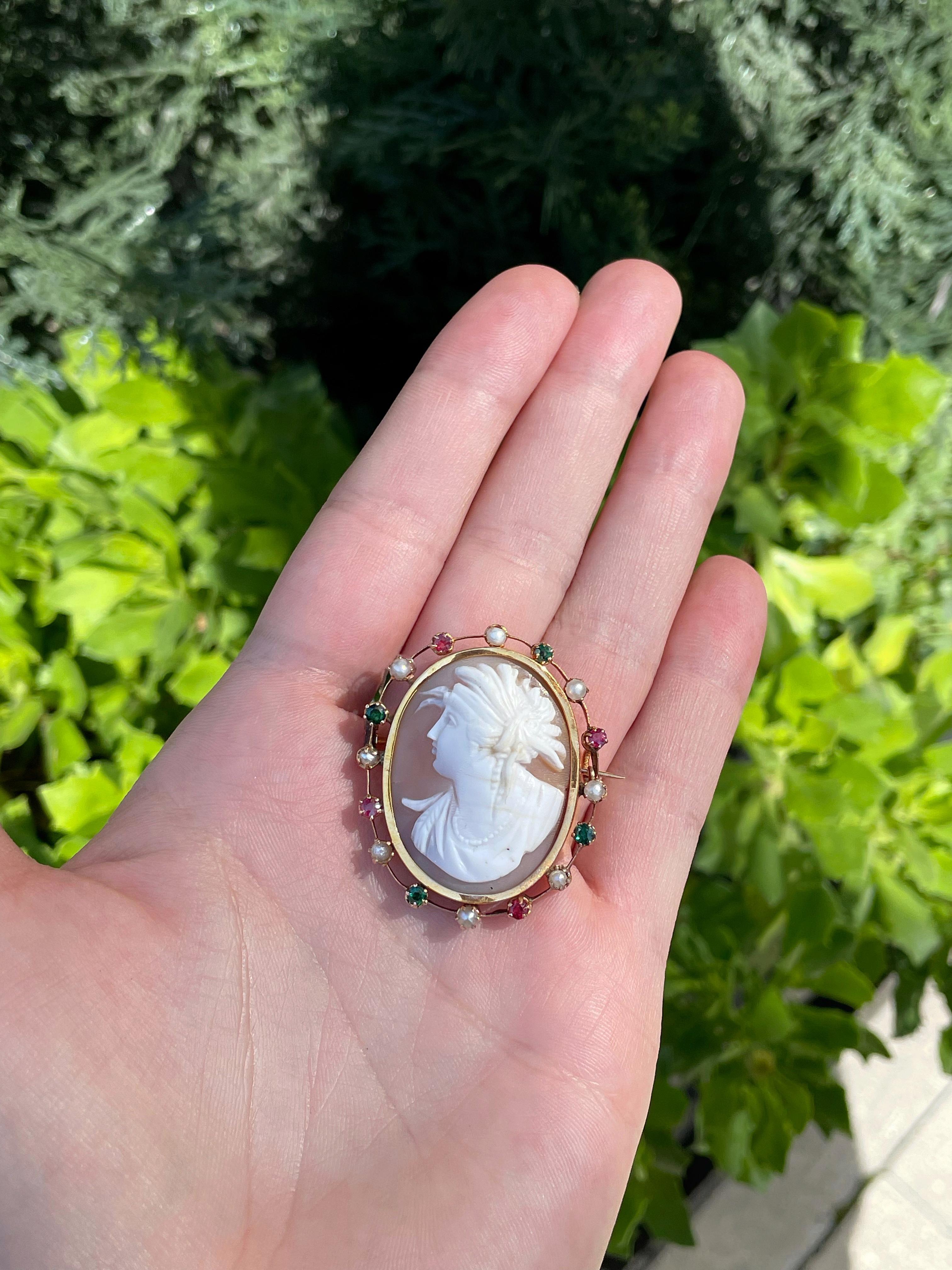 This is an elegant and rare Victorian pin brooch crafted in 14K yellow gold. The cameo is carved from shell. The frame is decorated with 4 red garnets (~0.32ct), 4 green glass and 8 cultured pearls. The inner part has a metal reinforcements.

The
