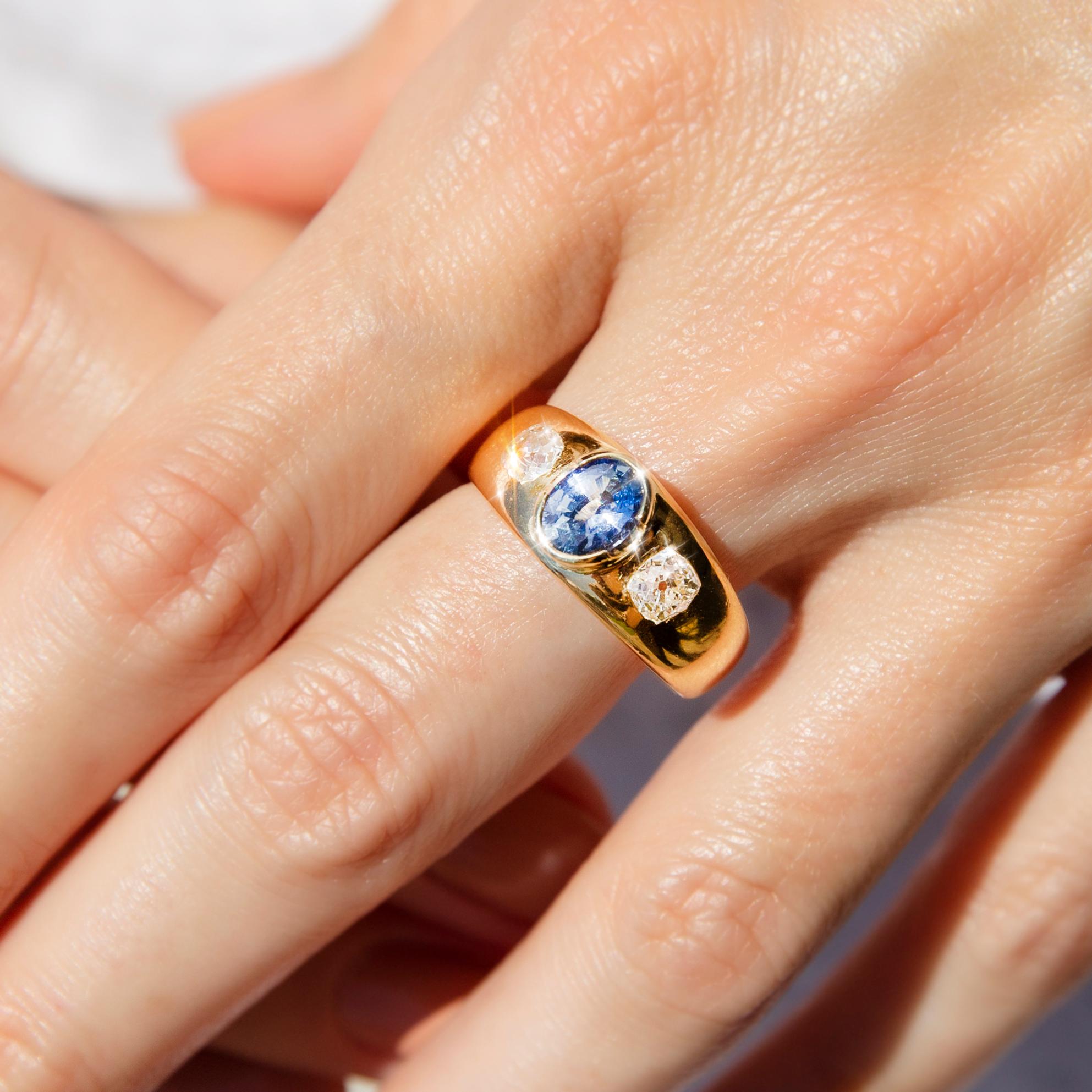 Forged in gleaming 18 carat yellow gold, this gorgeous Victorian-era trilogy ring features a darling oval mid-blue Ceylon-type sapphire in an elegant partial rubover setting between a duo of shimmering Old Mine Cut diamonds. This antique splendour