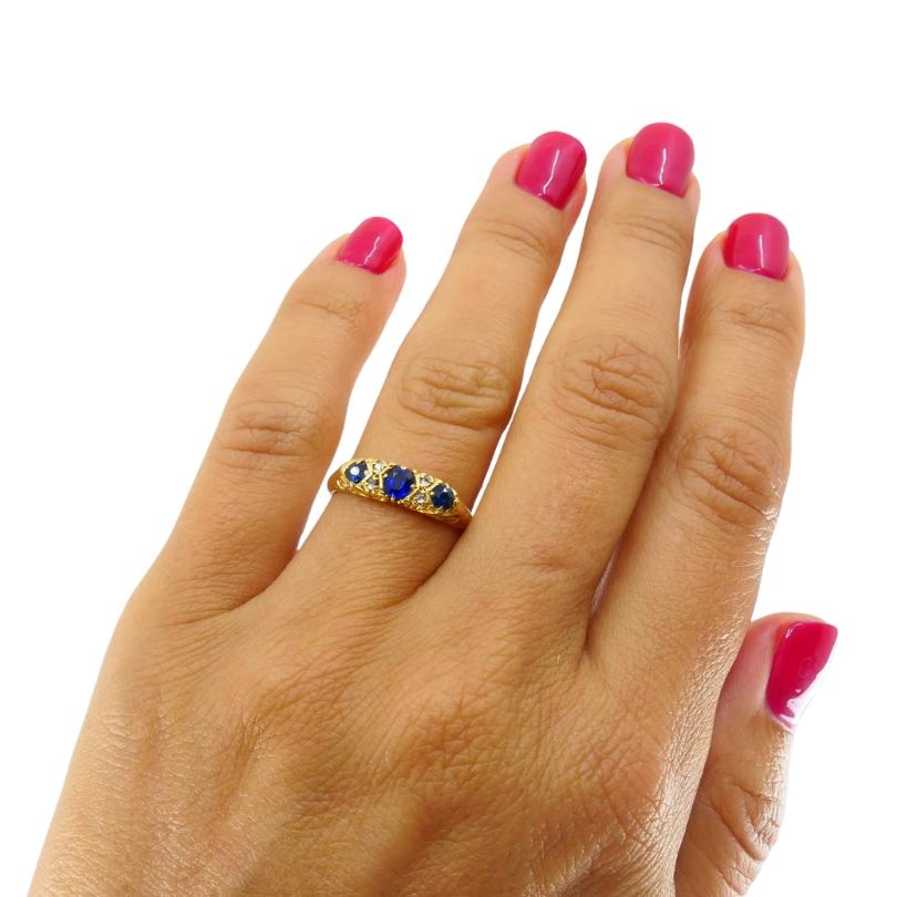 Un anello di epoca vittoriana splendidamente lavorato che mette in mostra un Trio di  zaffiri blu accentuati da delicati diamanti taglio vecchio, il tutto incastonato in un ornato oro giallo 18 carati. Le intricate volute e il design senza tempo